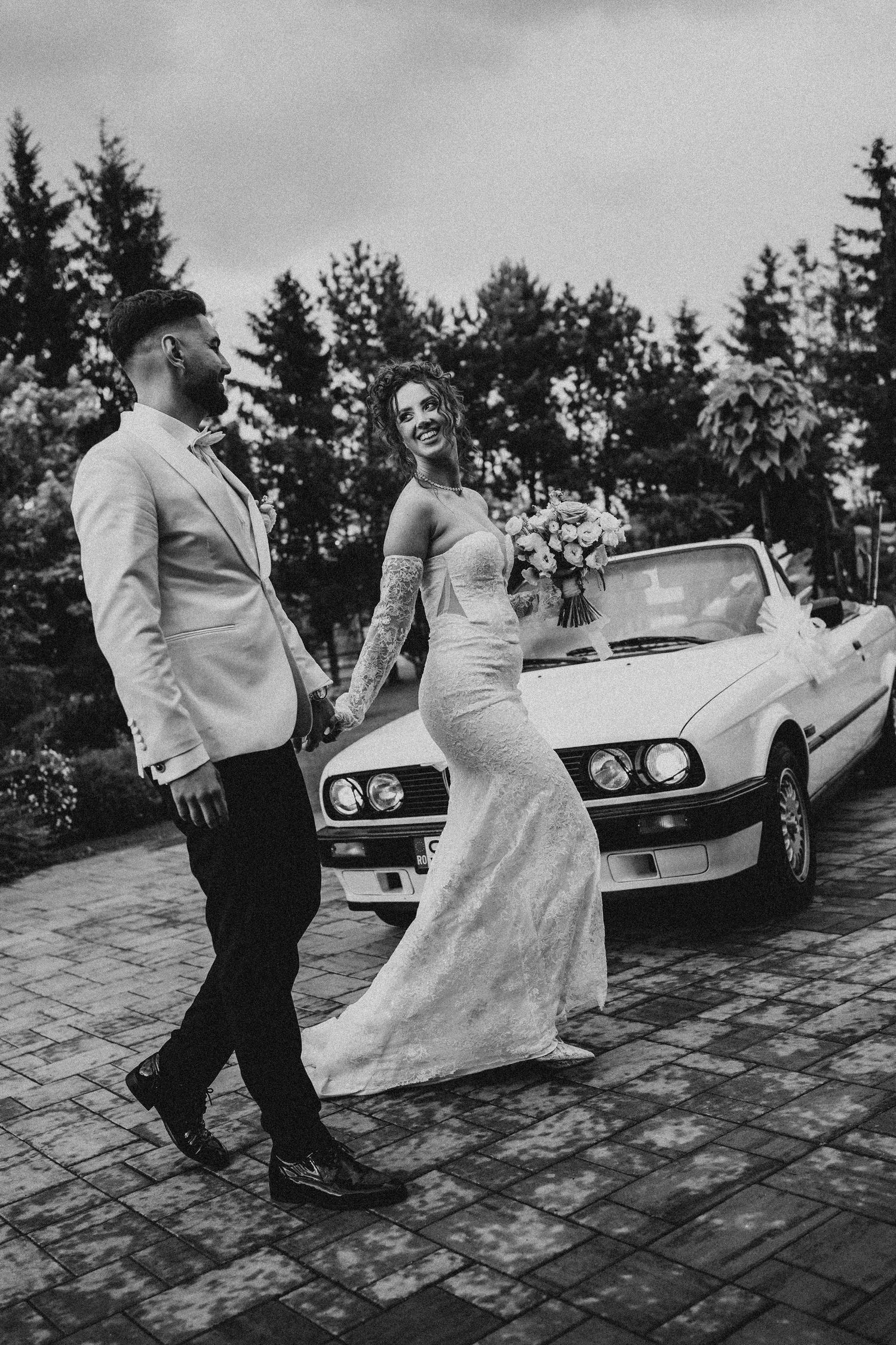 Black and white photo of a bride and groom holding hands beside a classic white convertible car on their wedding day.