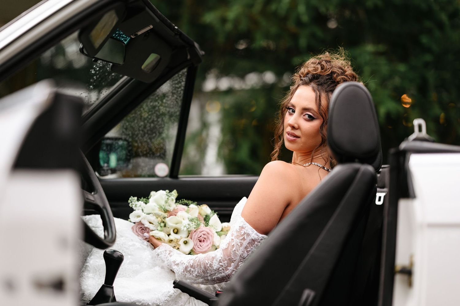 Bride in white lace gown holding bouquet of pink and white flowers, seated in a white convertible car.