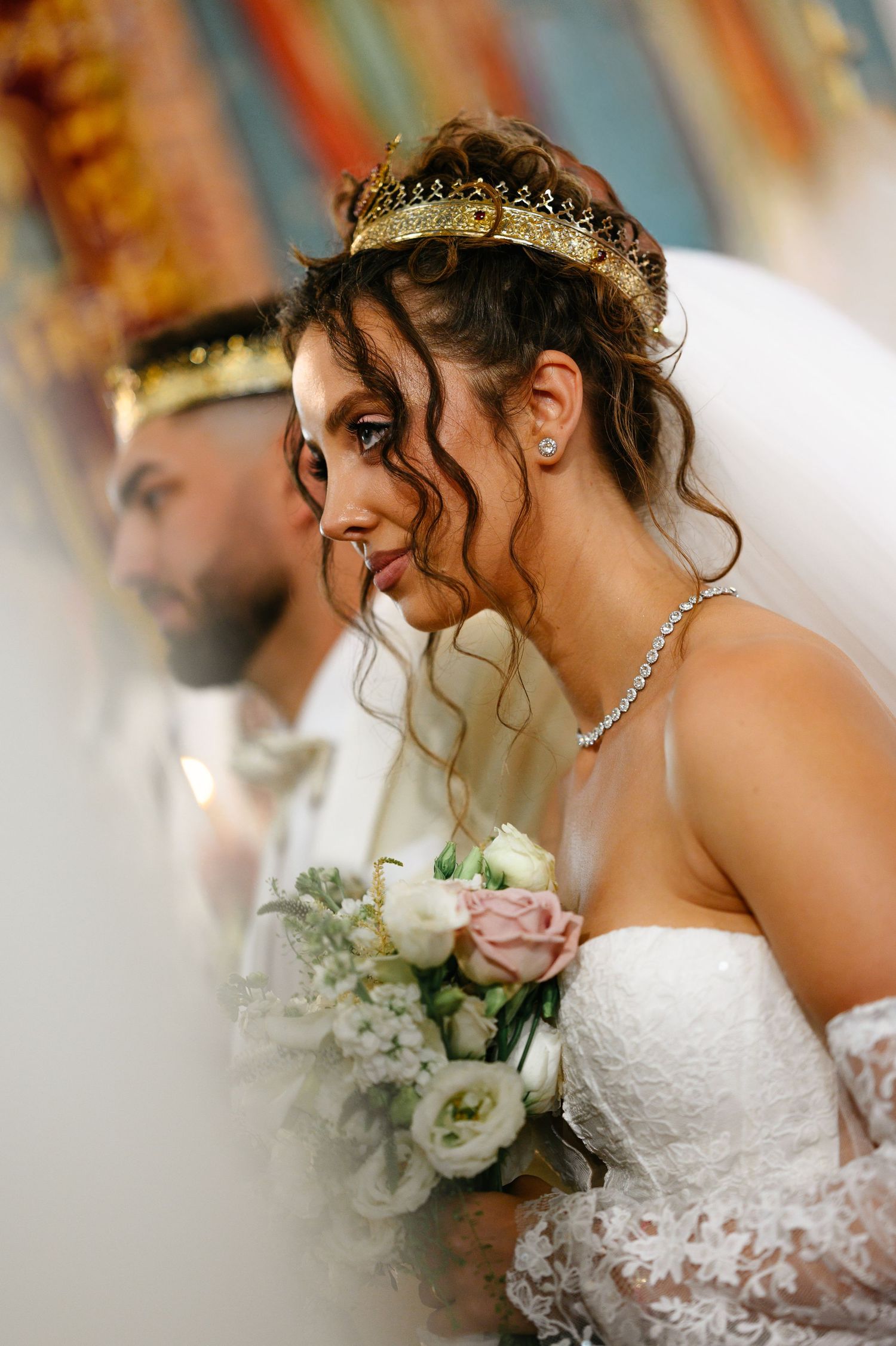Bride wearing gold crown tiara holds white and pink bouquet beside groom during wedding ceremony.