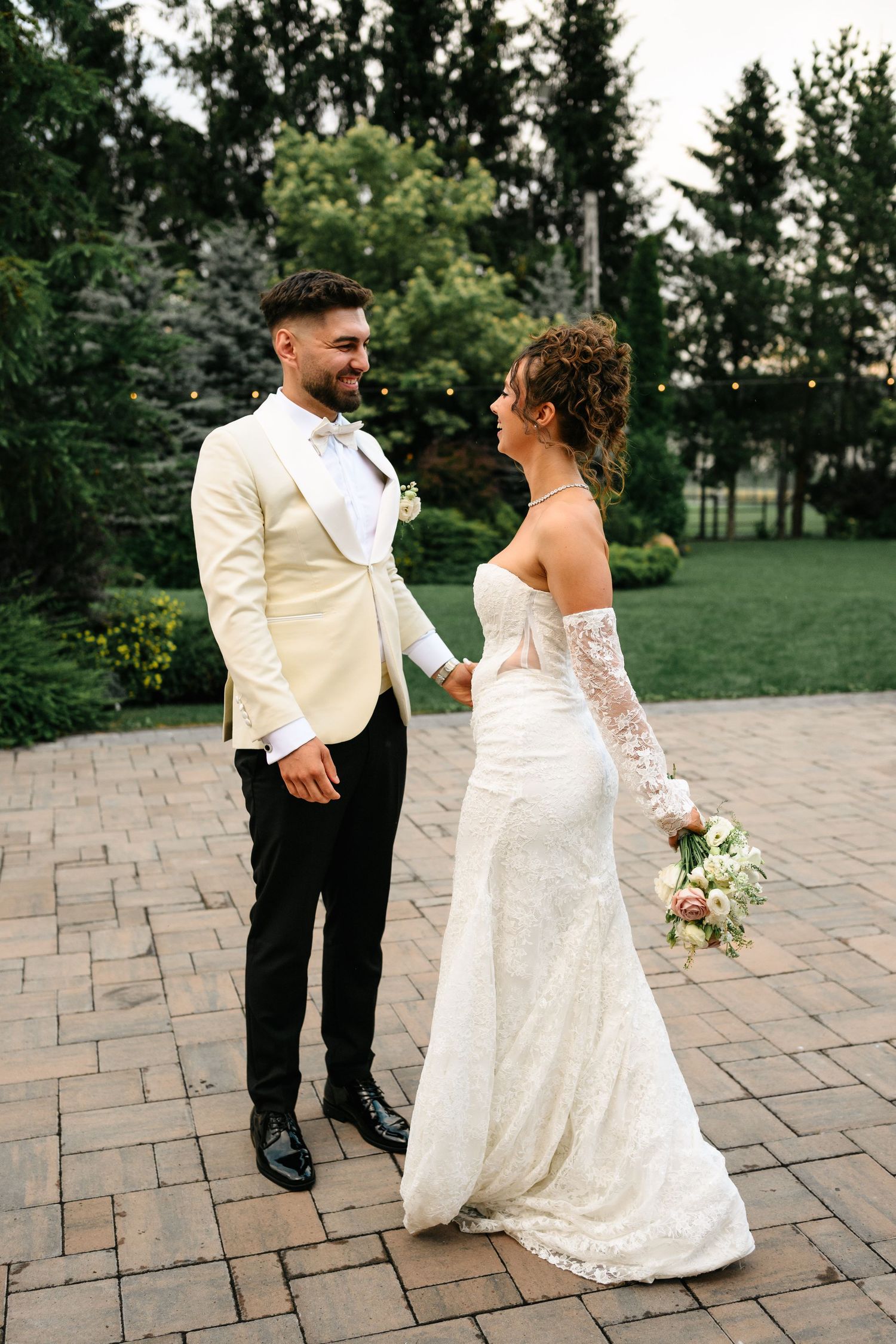 A bride in a lace gown holds a bouquet while facing her groom in a cream tuxedo on a brick patio.