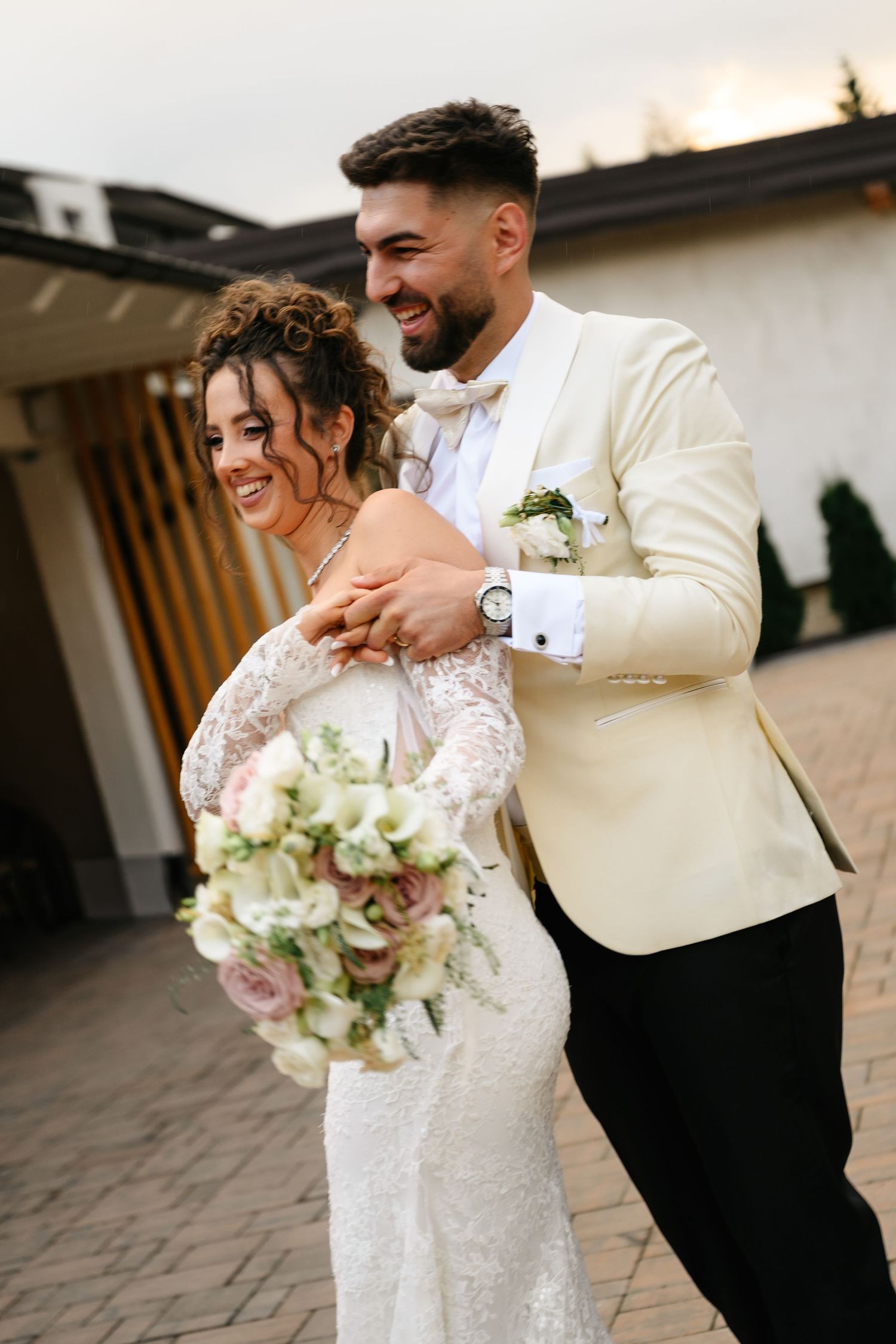 Happy newlyweds embrace outdoors, bride holding floral bouquet in lace gown, groom in cream tuxedo jacket.