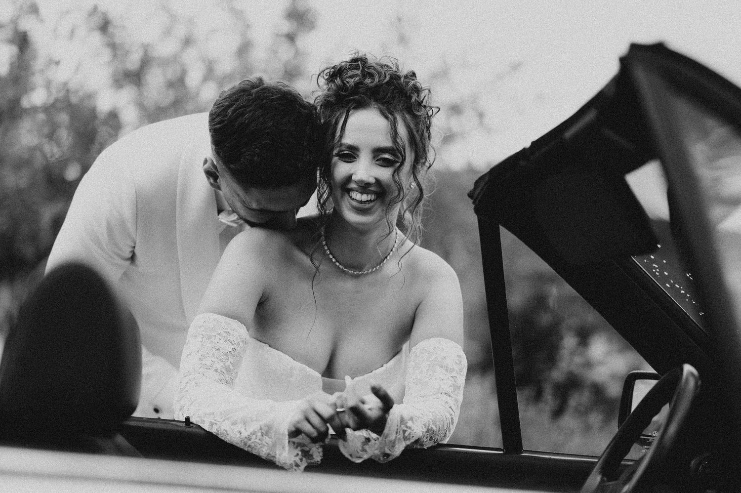 Black and white photo of a smiling bride and groom sharing a romantic moment in a vintage convertible car.