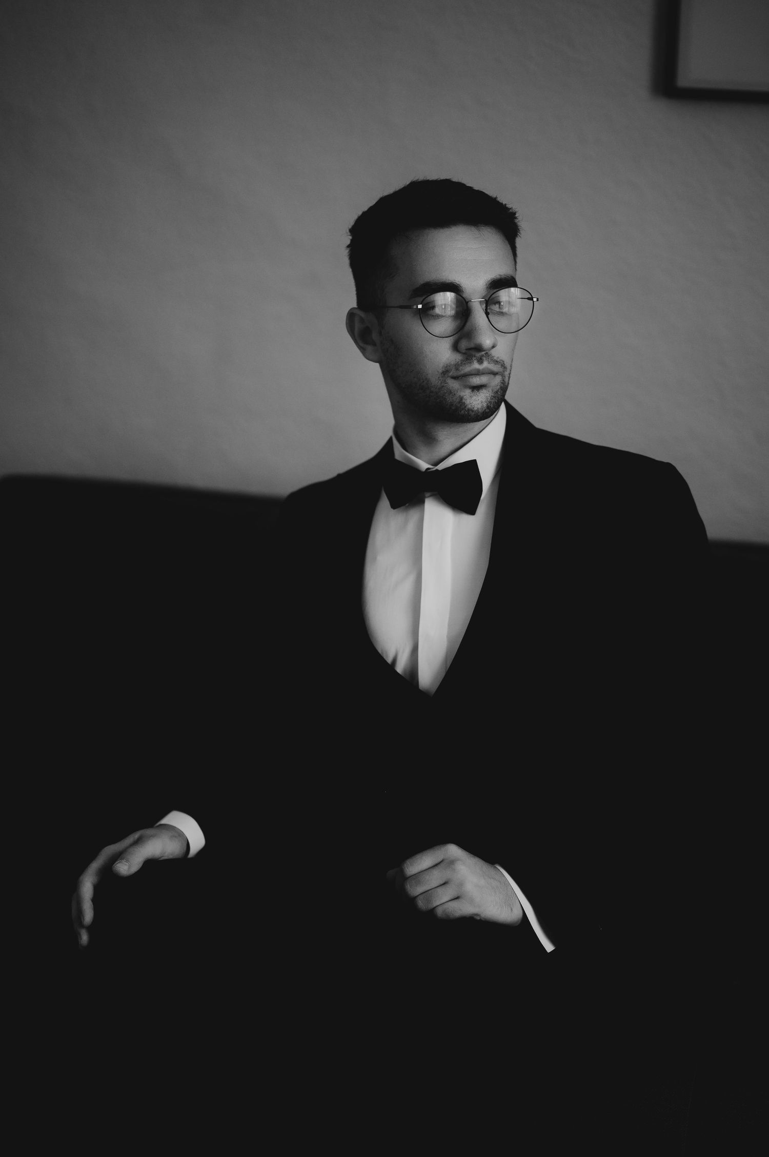 Stylish groom in black tuxedo adjusting jacket, wearing round glasses in moody black and white photo.
