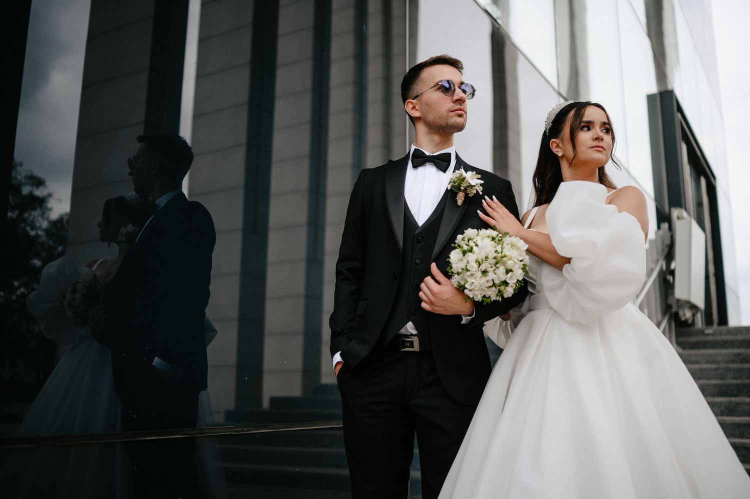 Elegant bride and groom posing on steps before tall columns, groom in tuxedo with sunglasses.