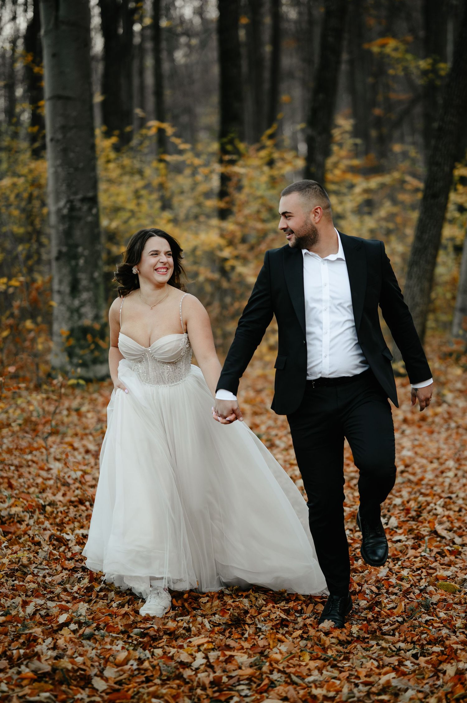 Bride and groom holding hands, walking through autumn forest with fallen leaves, smiling at each other in wedding attire.