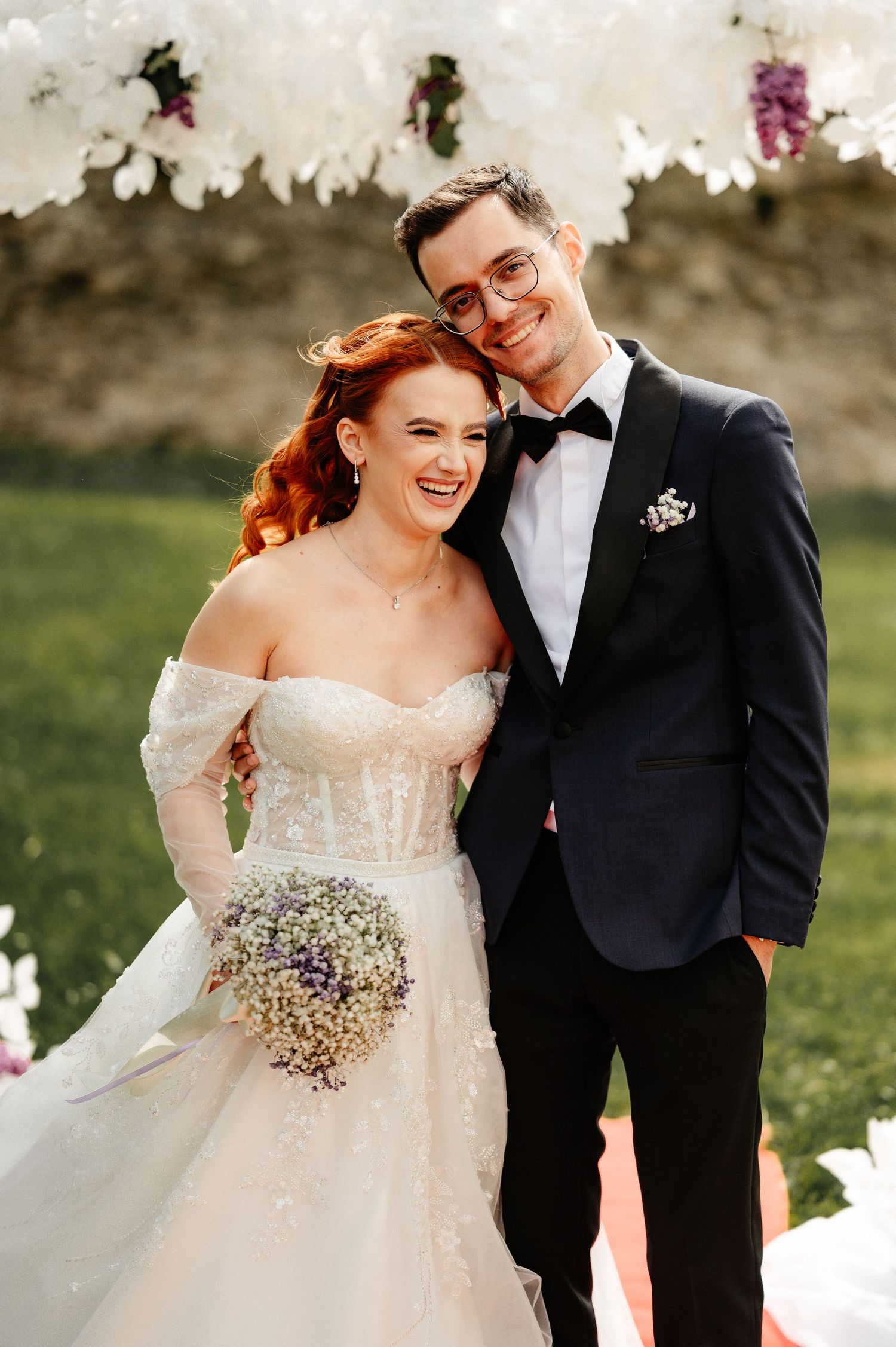 A smiling bride in a white off-shoulder gown holds a lavender bouquet beside a groom in a navy tuxedo at an outdoor wedding.