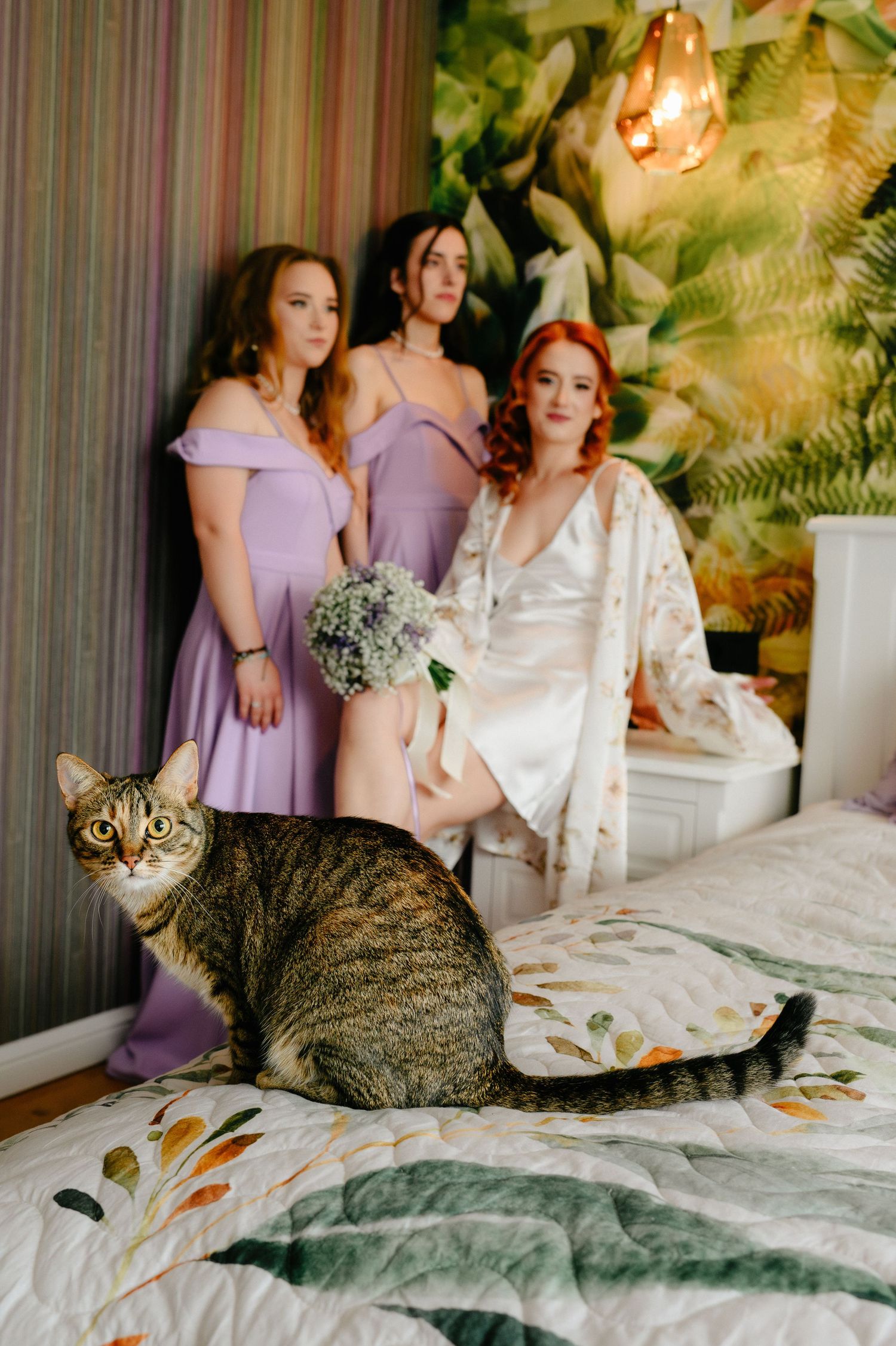Bride in white robe holds bouquet with bridesmaids in purple dresses and a tabby cat on a floral bed.