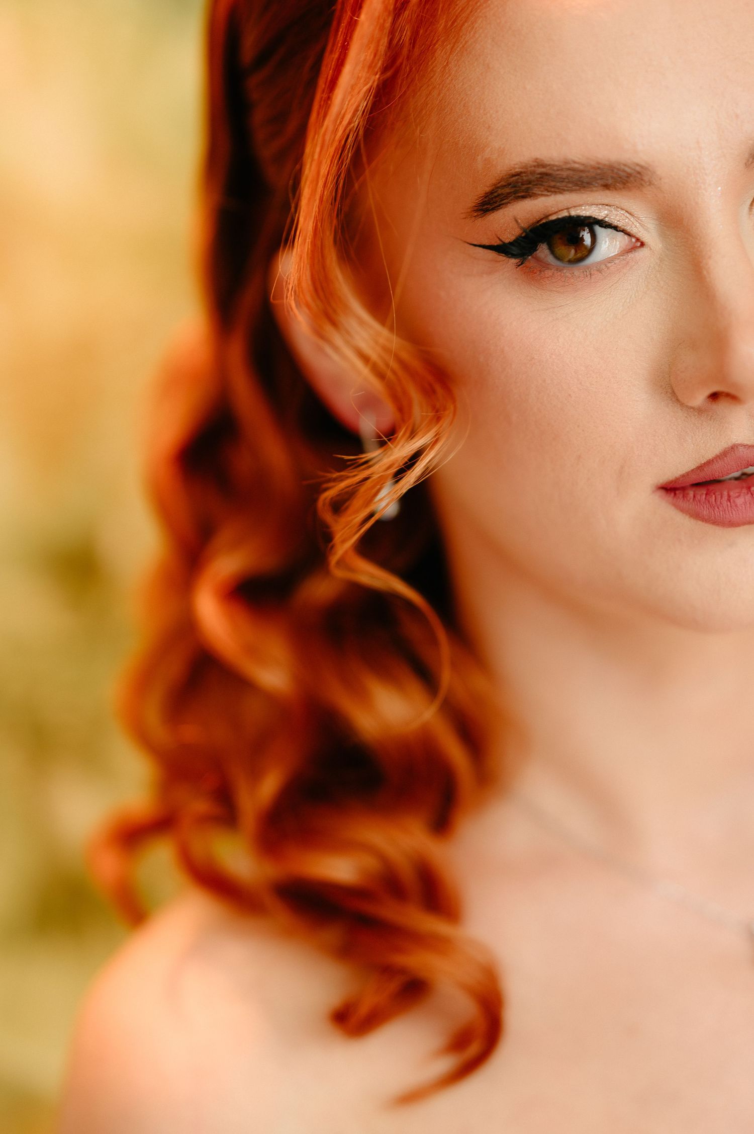 Close-up portrait of a young woman with curly red hair, cat-eye liner, and subtle pink lips against a golden background.