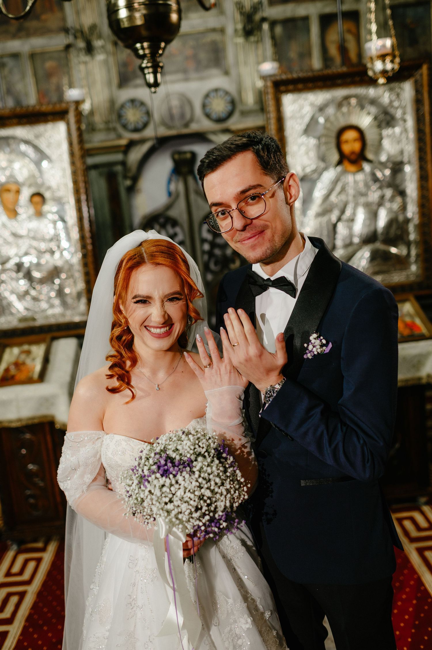 Newlywed couple smiling in Orthodox church, bride holding baby's breath bouquet, groom in navy suit showing wedding rings.