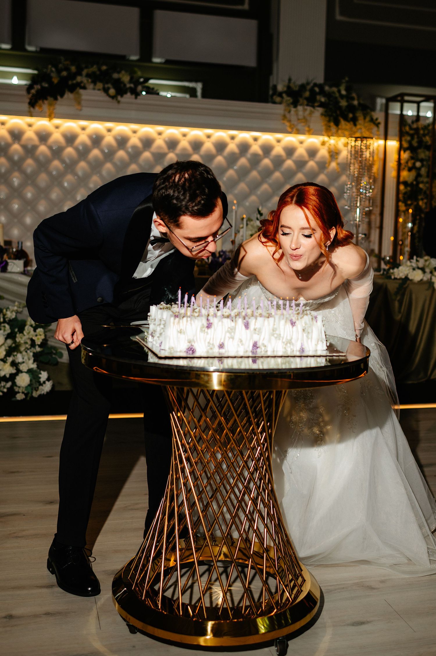 Bride and groom blowing out candles on their wedding cake at an elegant reception venue.