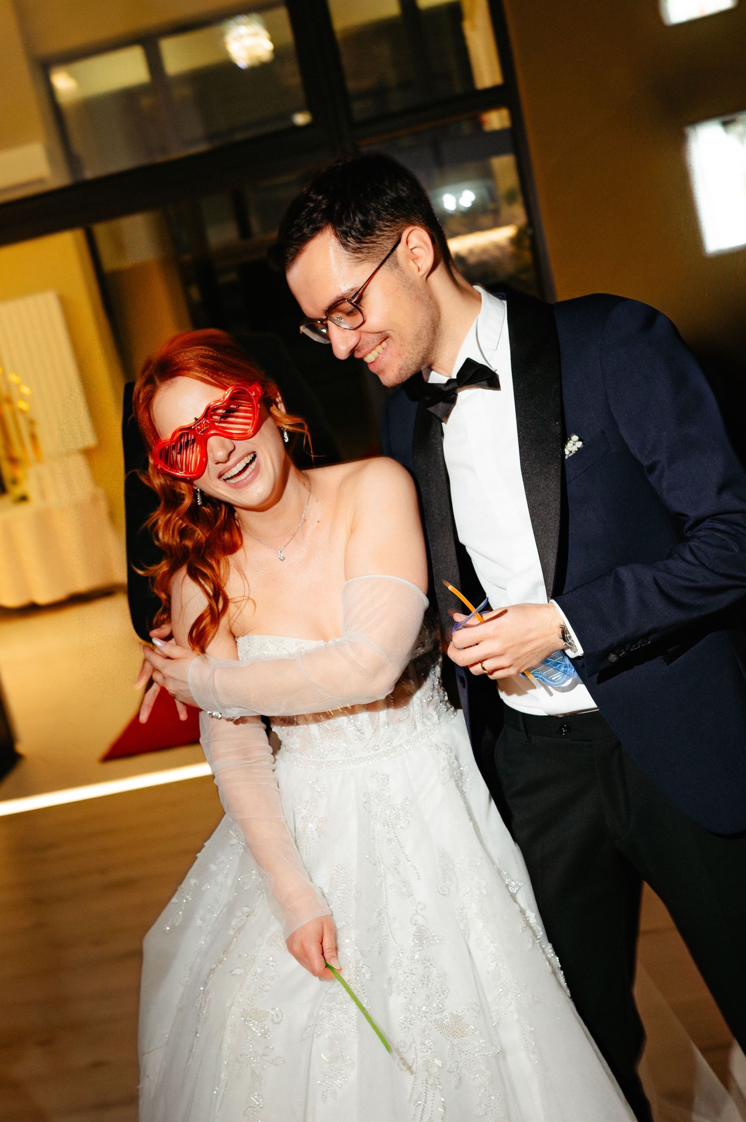 Bride in white gown with red novelty glasses laughs with groom in navy tuxedo at wedding reception.