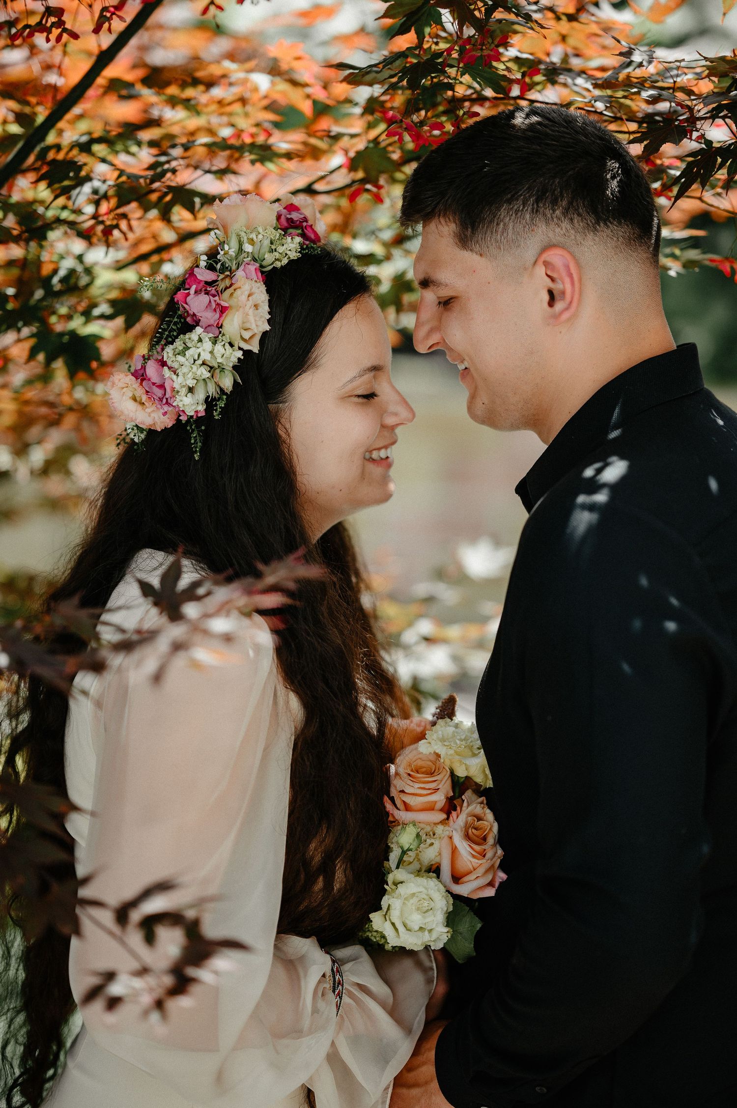 Smiling bride with floral crown and groom touching foreheads under autumn maple tree.