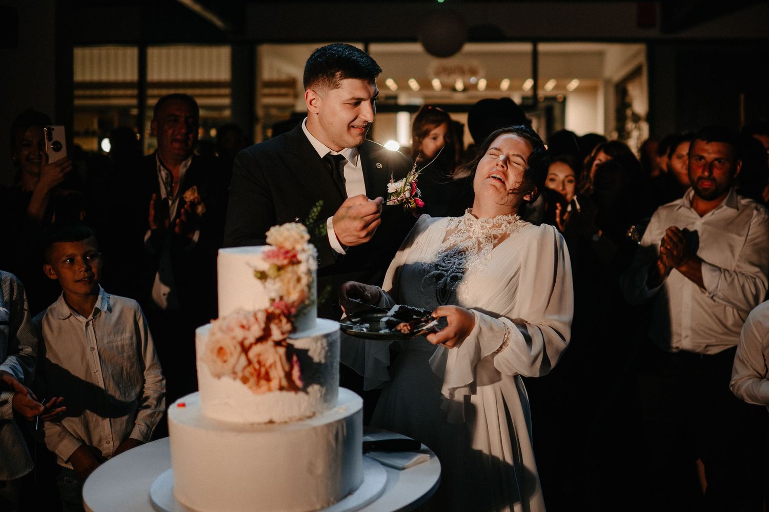 Laughing newlyweds cutting a white wedding cake with floral decor at their reception.