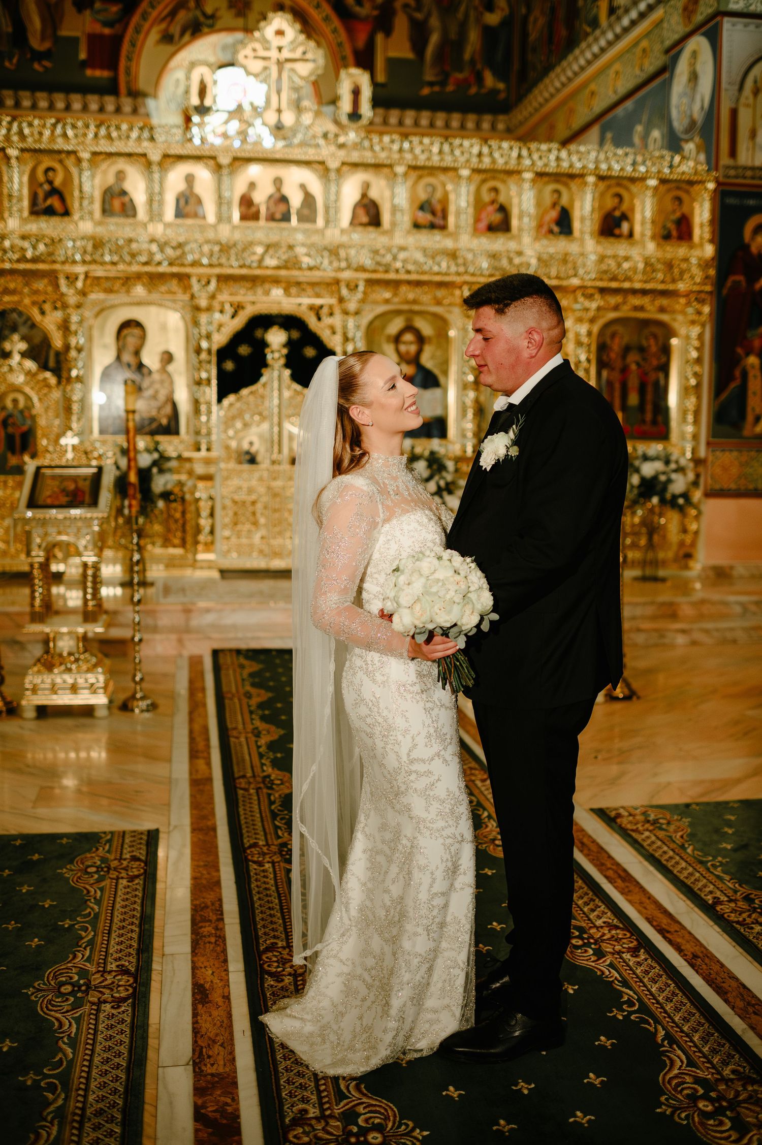 Bride and groom smiling at each other inside ornate Orthodox church.