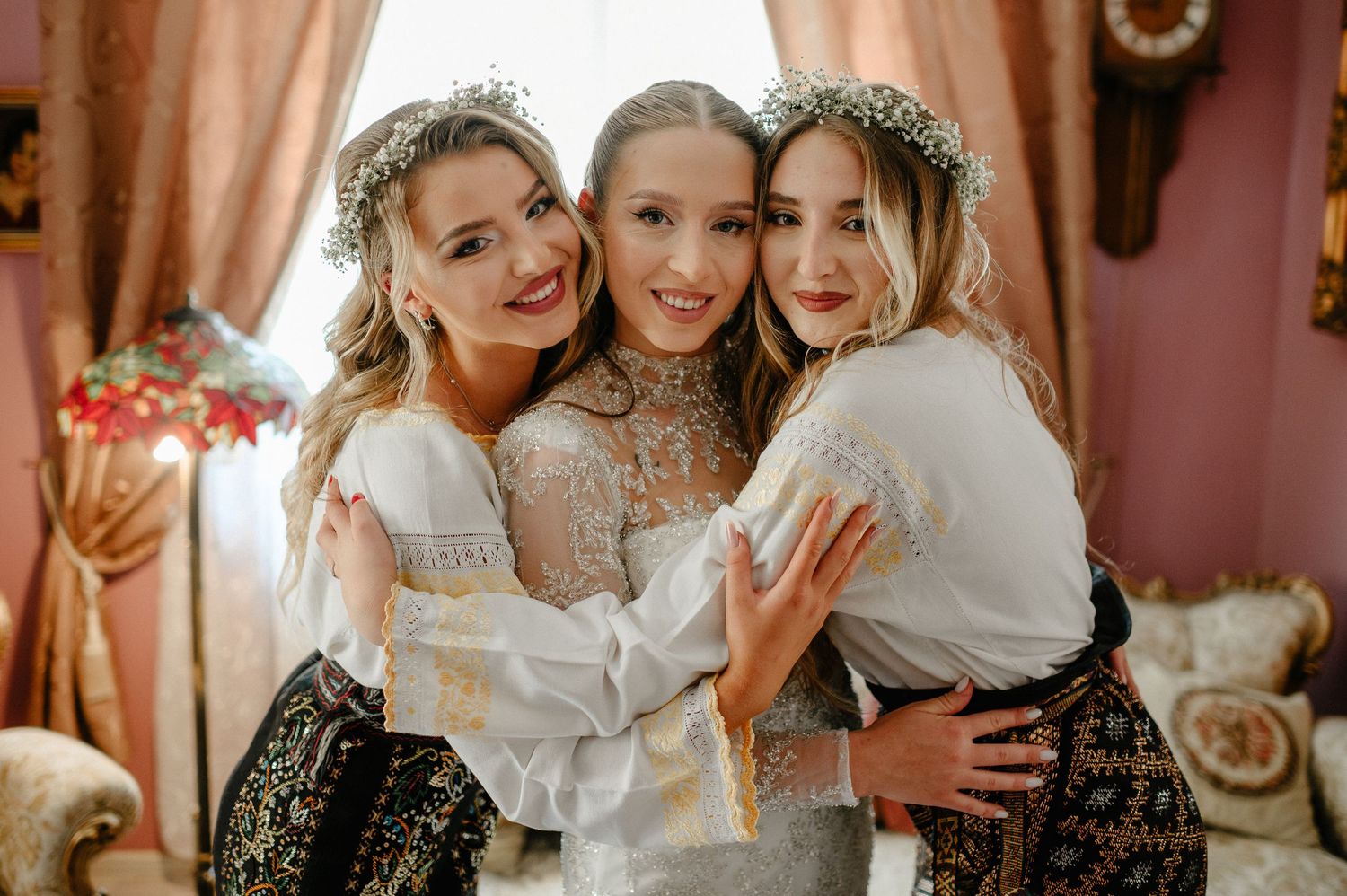 Three women in traditional white embroidered dresses with floral crowns embracing and smiling together.