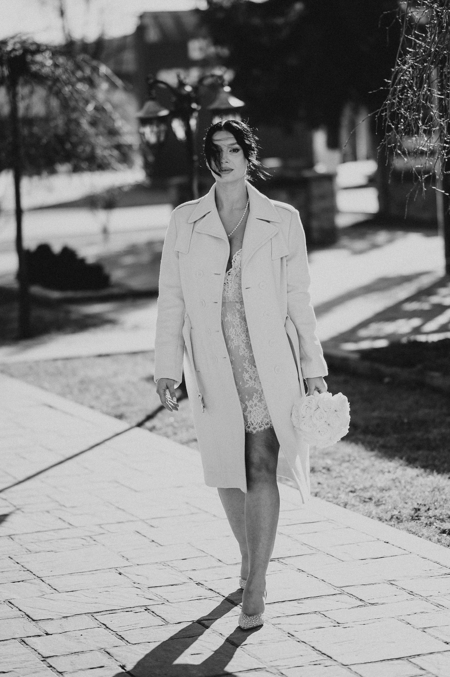 Elegant bride in white coat holding bouquet walks on stone path in black and white photo.