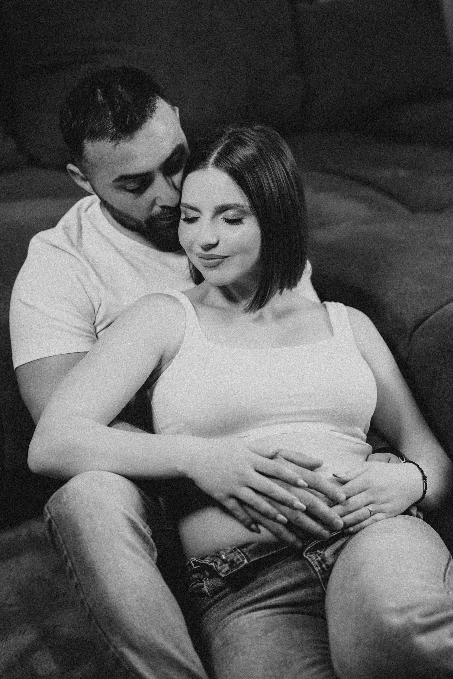 Black and white photo of a couple sitting together, man embracing pregnant woman from behind on a couch.