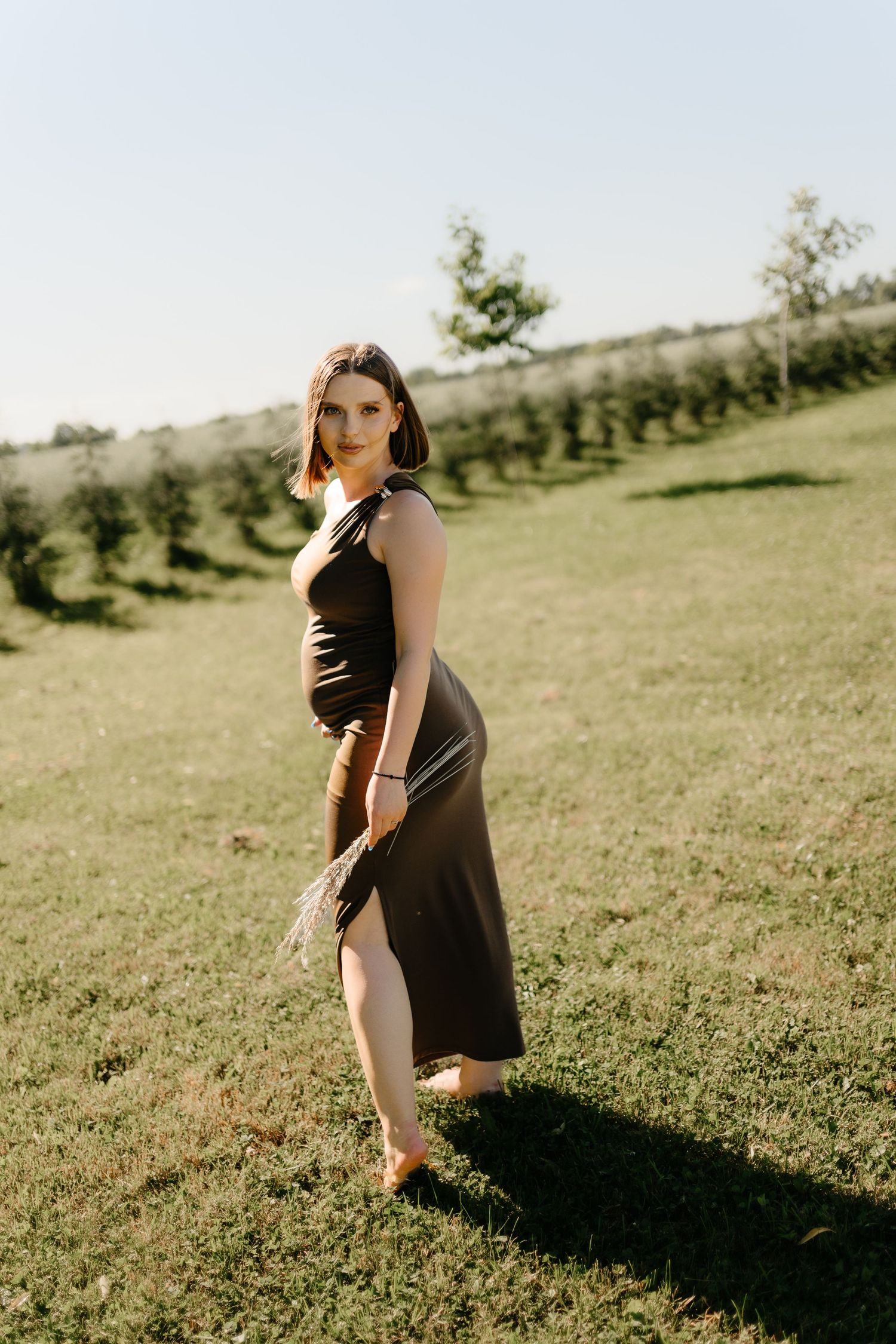 Pregnant woman in elegant dark dress posing in sunlit green orchard field.