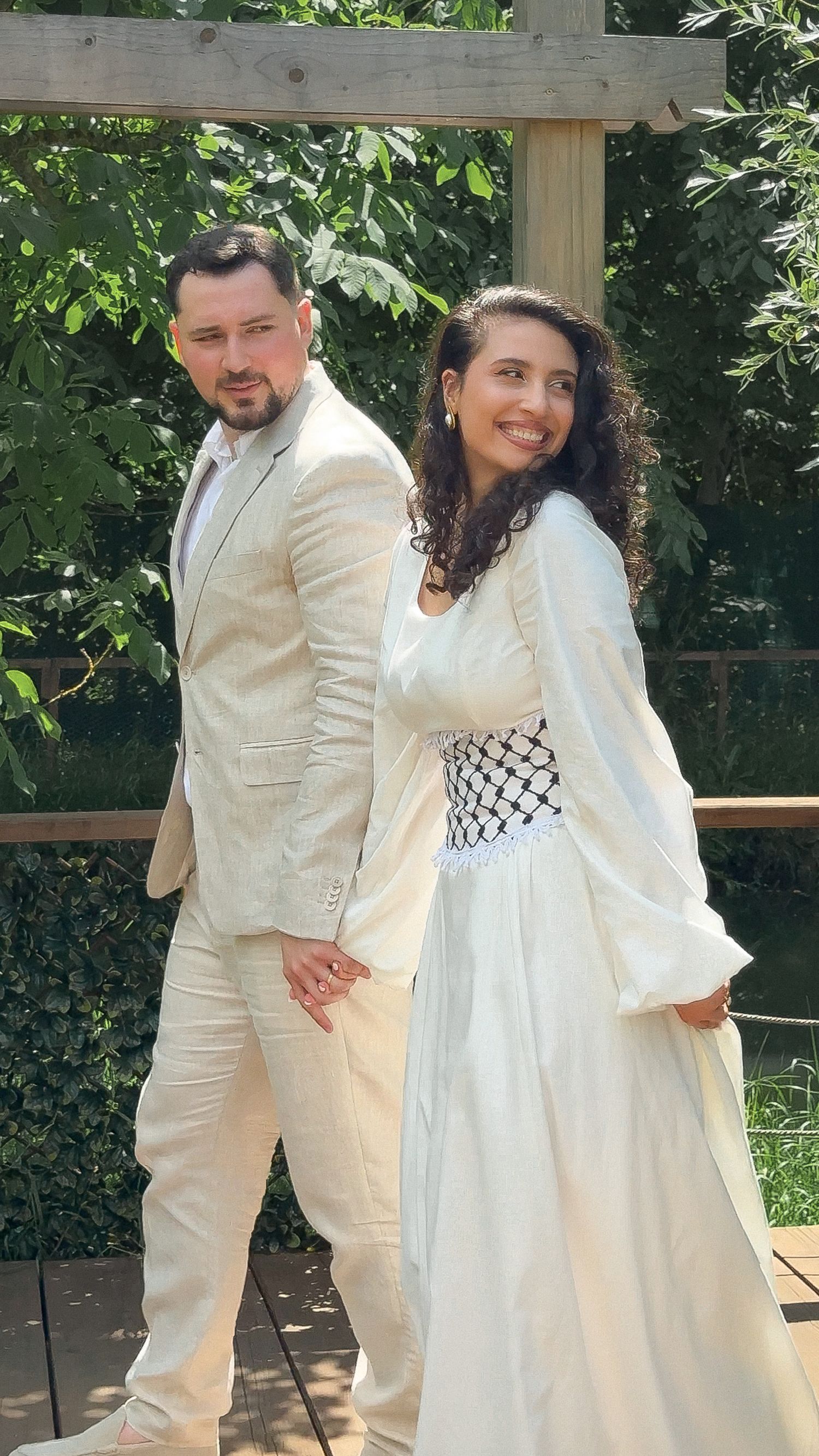 Bride in white gown with keffiyeh corset and groom in beige suit hold hands outdoors at garden wedding.