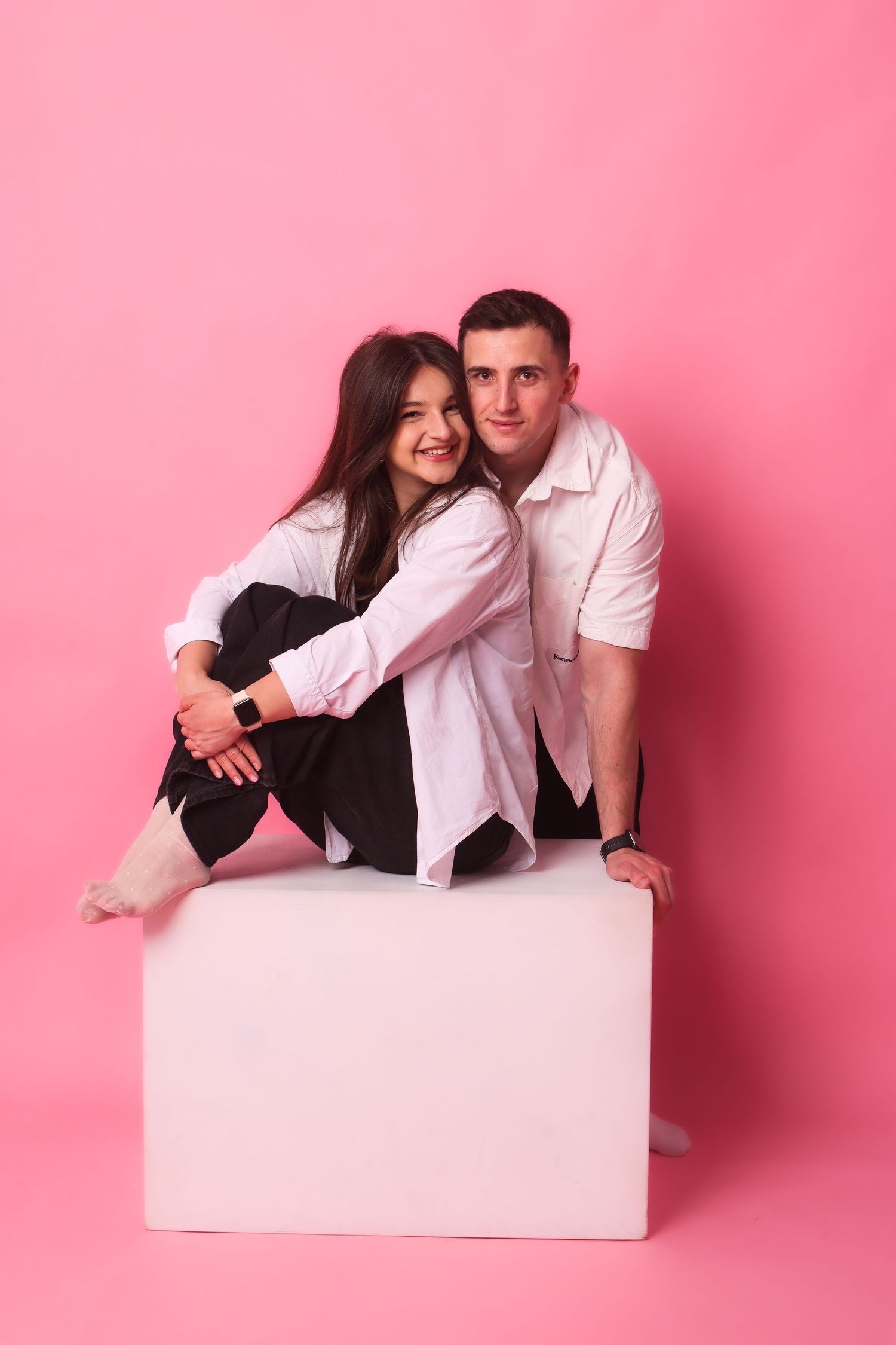 A smiling couple in black and white outfits sit together on a white cube against pink background.