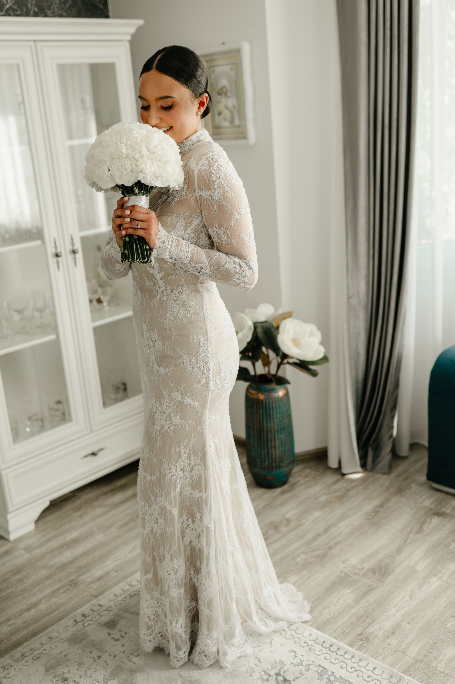 Bride in fitted long sleeve lace gown smelling white hydrangea bouquet indoors.