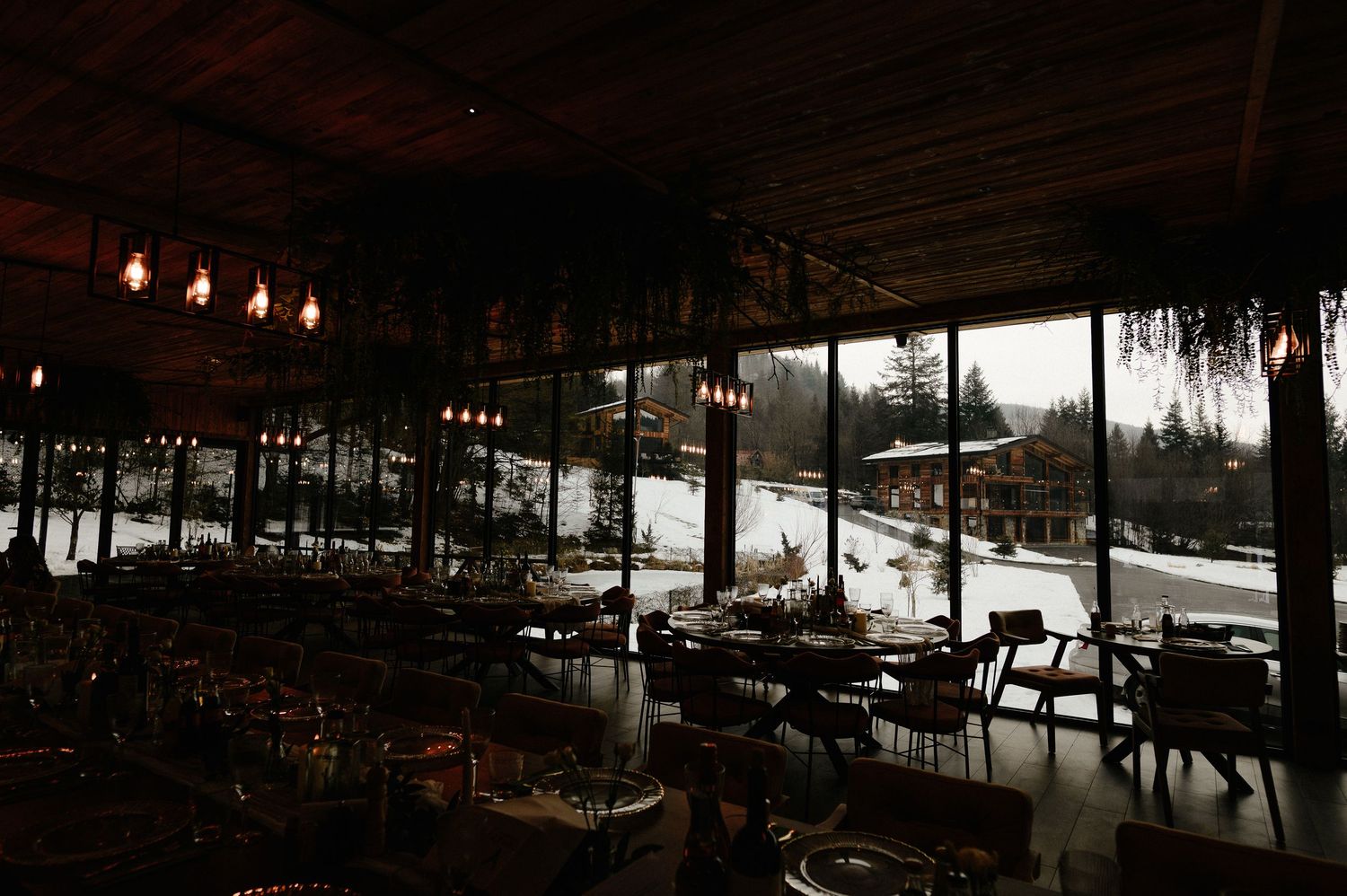 Dimly lit rustic restaurant interior with large windows overlooking snowy winter landscape at dusk.