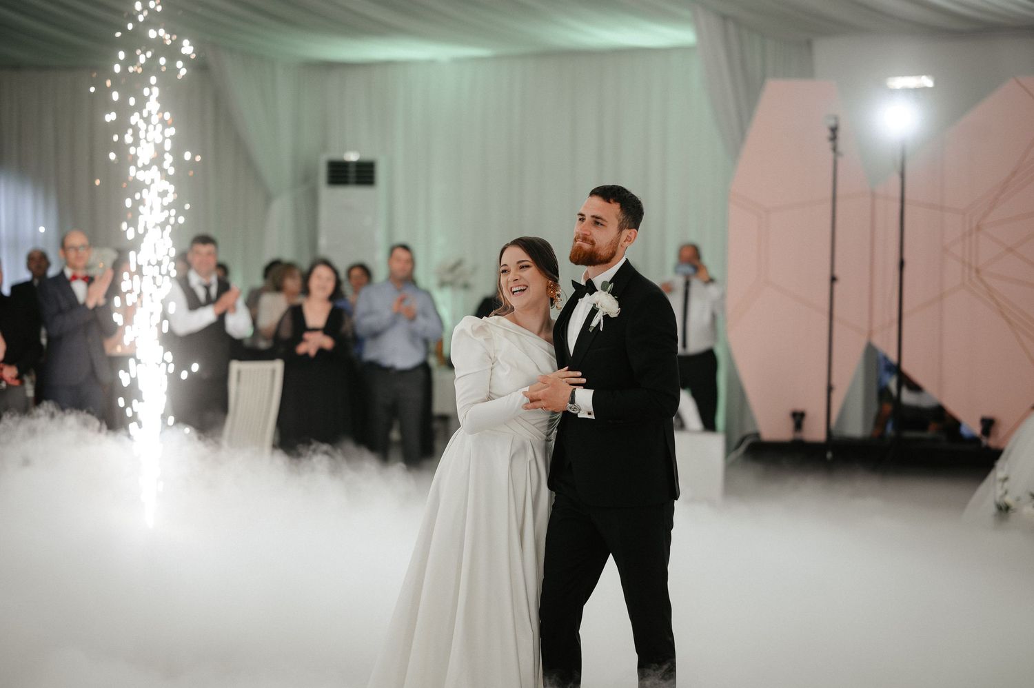 Bride and groom smile joyfully during first dance on fog-covered dance floor with sparkler effects.