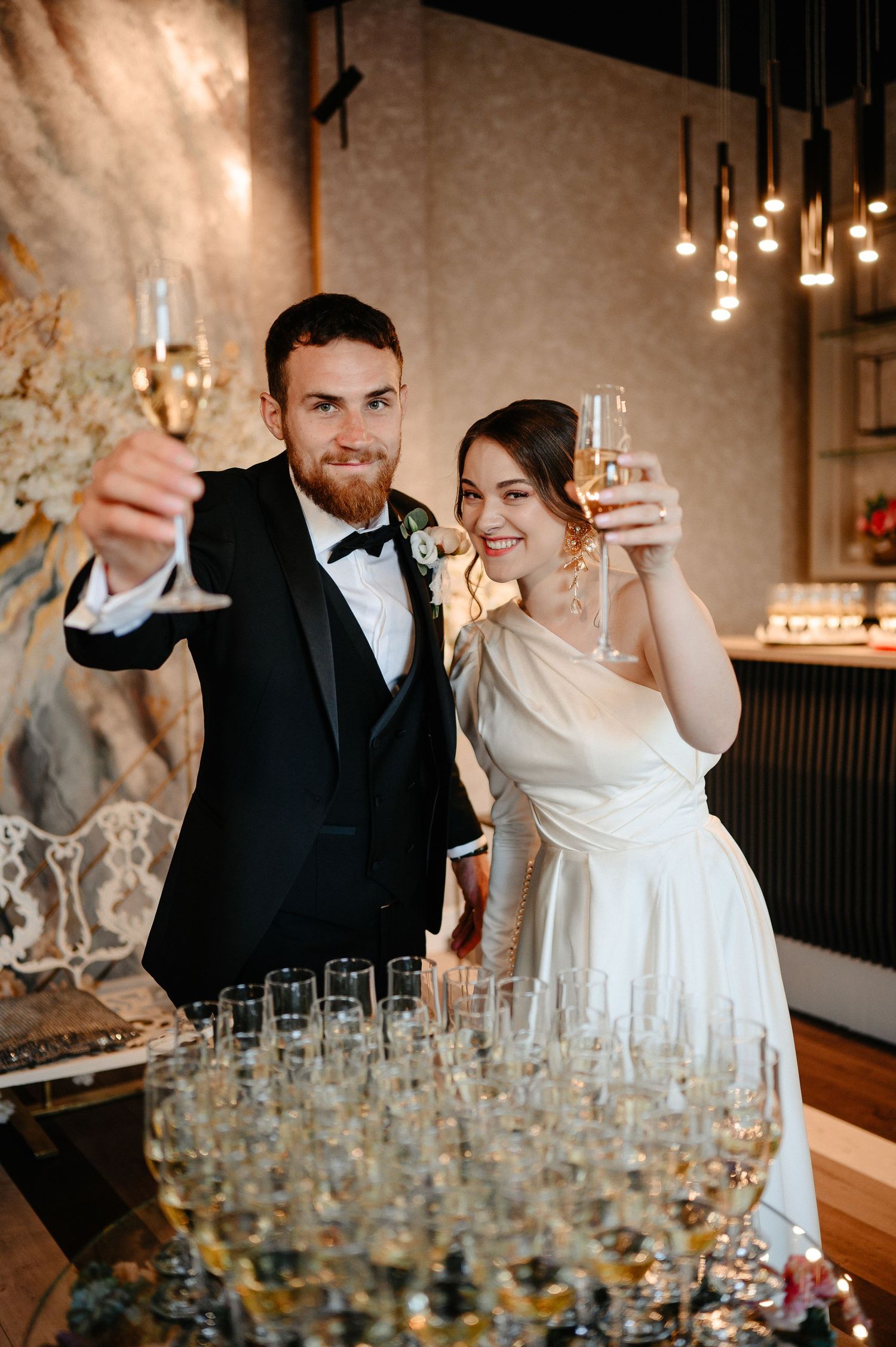 Newlyweds raising champagne glasses in toast beside pyramid of flutes at reception.