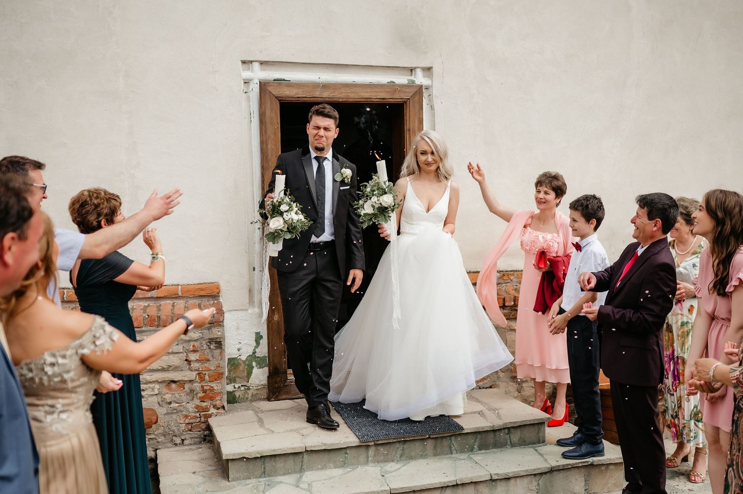Newlyweds exit a charming stone church as guests celebrate with applause and confetti.