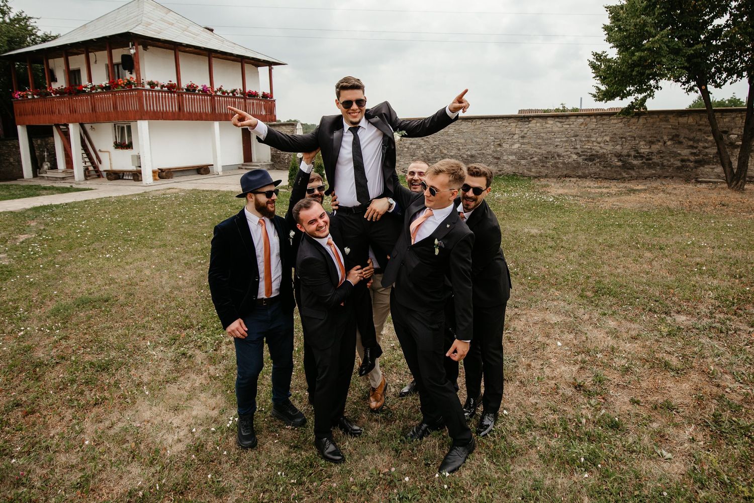 Groomsmen lifting the groom in celebration outdoors near a white building during a fun wedding party moment.