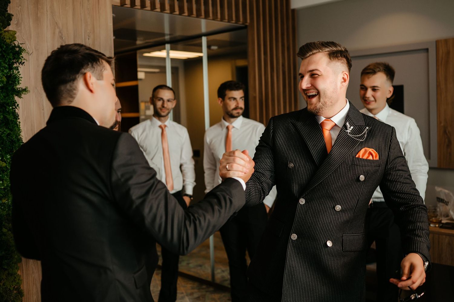 Men in formal suits shaking hands and laughing at a wedding celebration gathering.