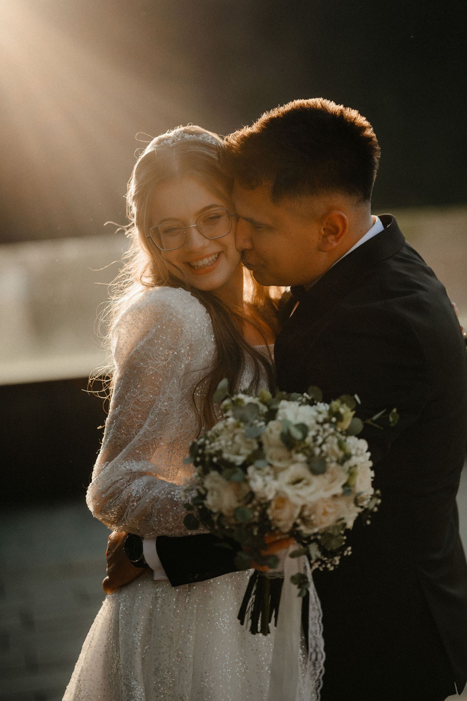 Groom embracing smiling bride in white lace gown holding white bouquet in warm sunlight.