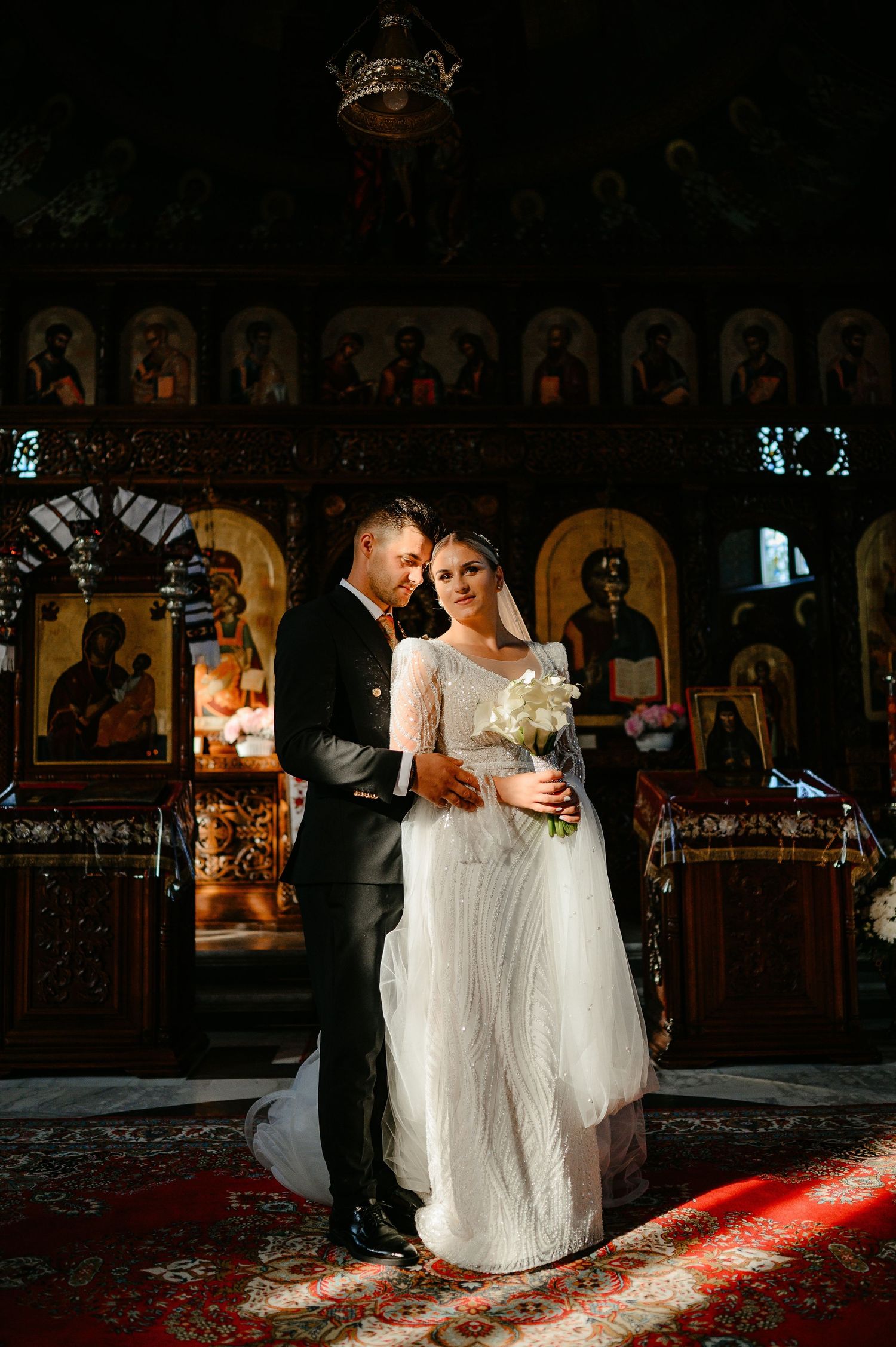 Bride and groom embracing inside an Orthodox church adorned with golden icons during their wedding ceremony.