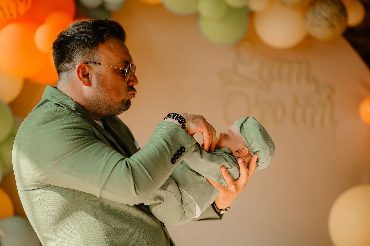 Father in sage green outfit carefully holding tiny newborn baby at colorful balloon celebration.