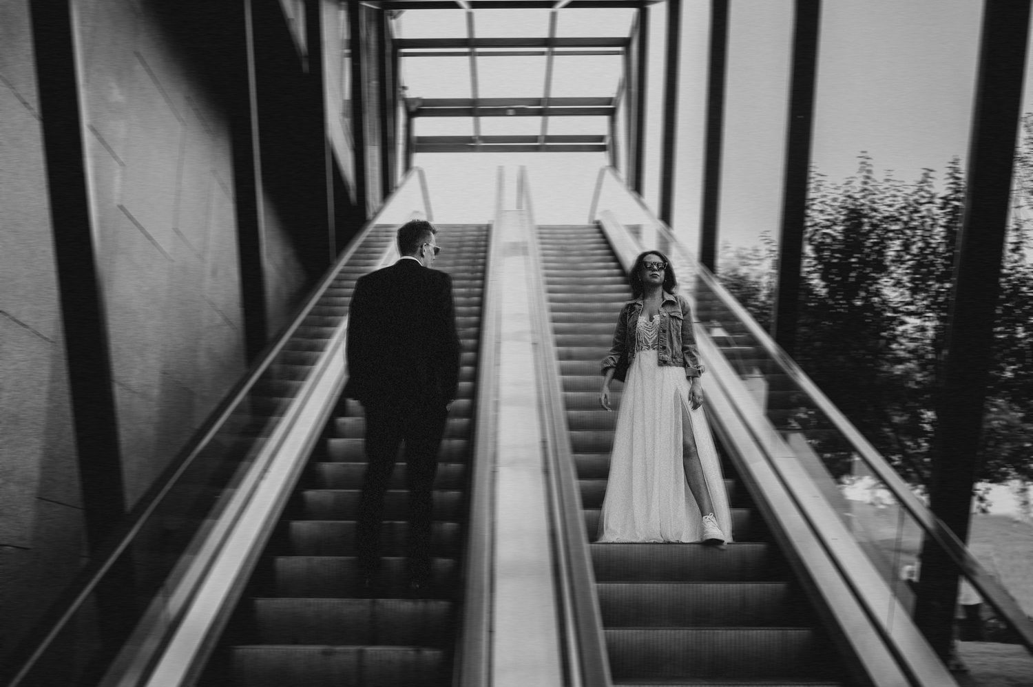 Bride and groom ride separate escalators facing each other in a modern glass building in black and white.