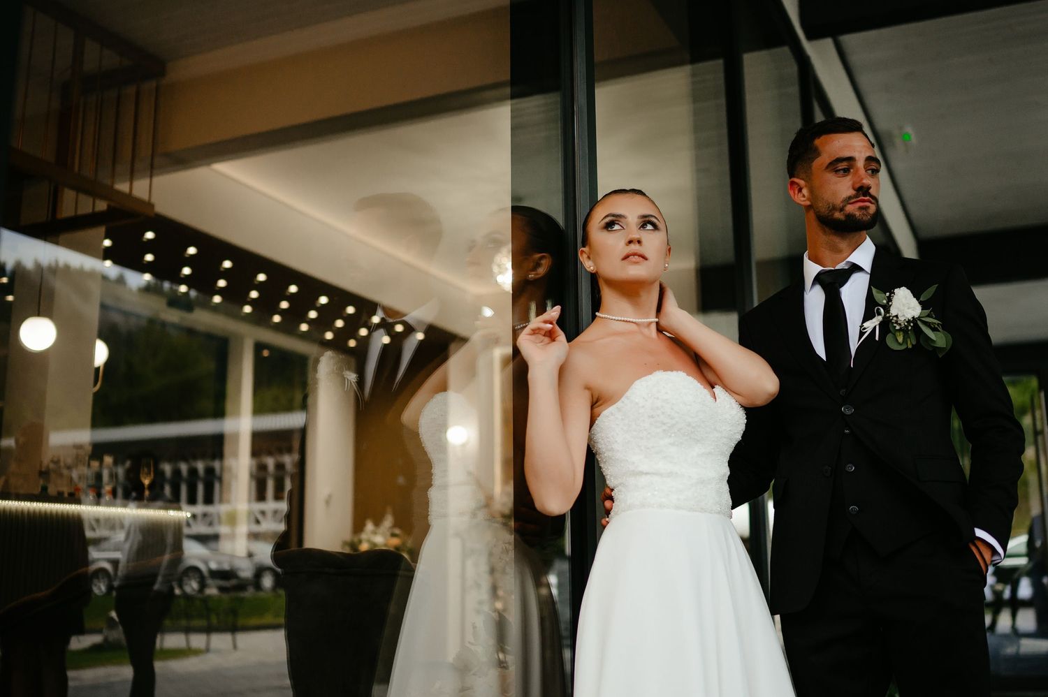 Stylish bride and groom posing against glass building exterior, bride touching necklace elegantly.