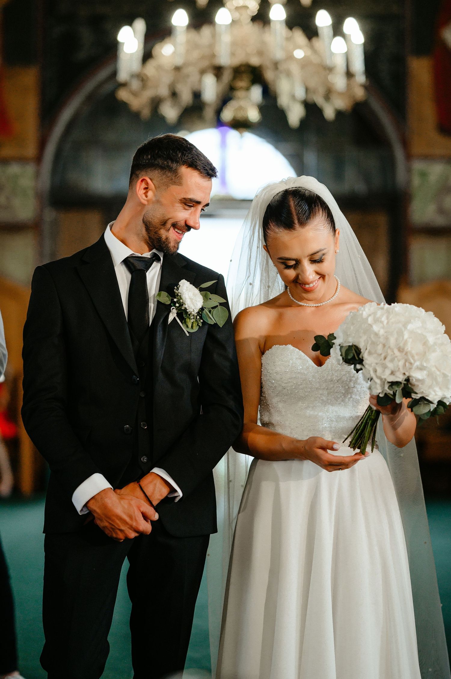 Smiling bride and groom share joyful moment during Orthodox church wedding ceremony under chandelier.