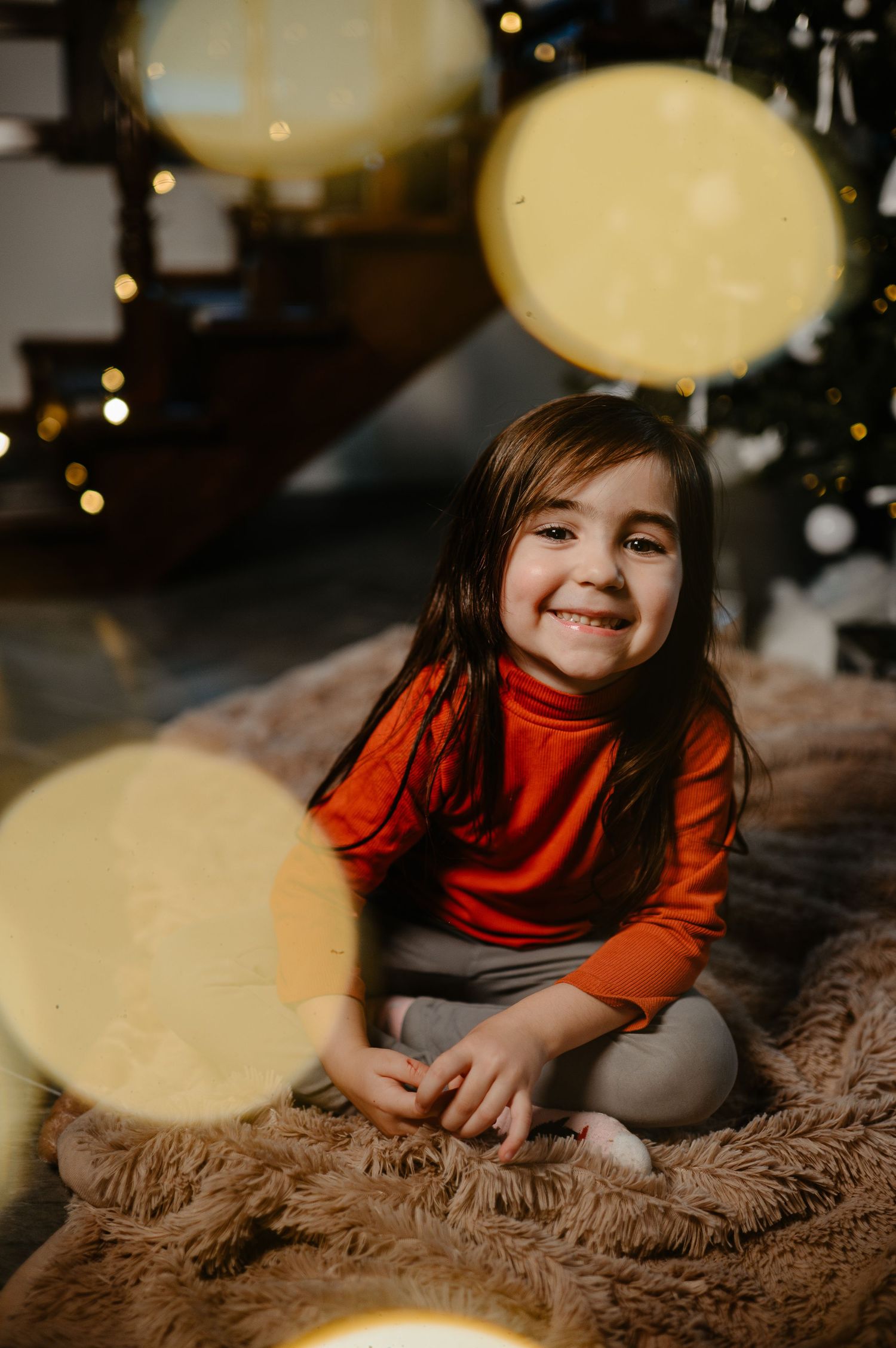 Happy little girl in red turtleneck sitting on fur rug near Christmas tree.