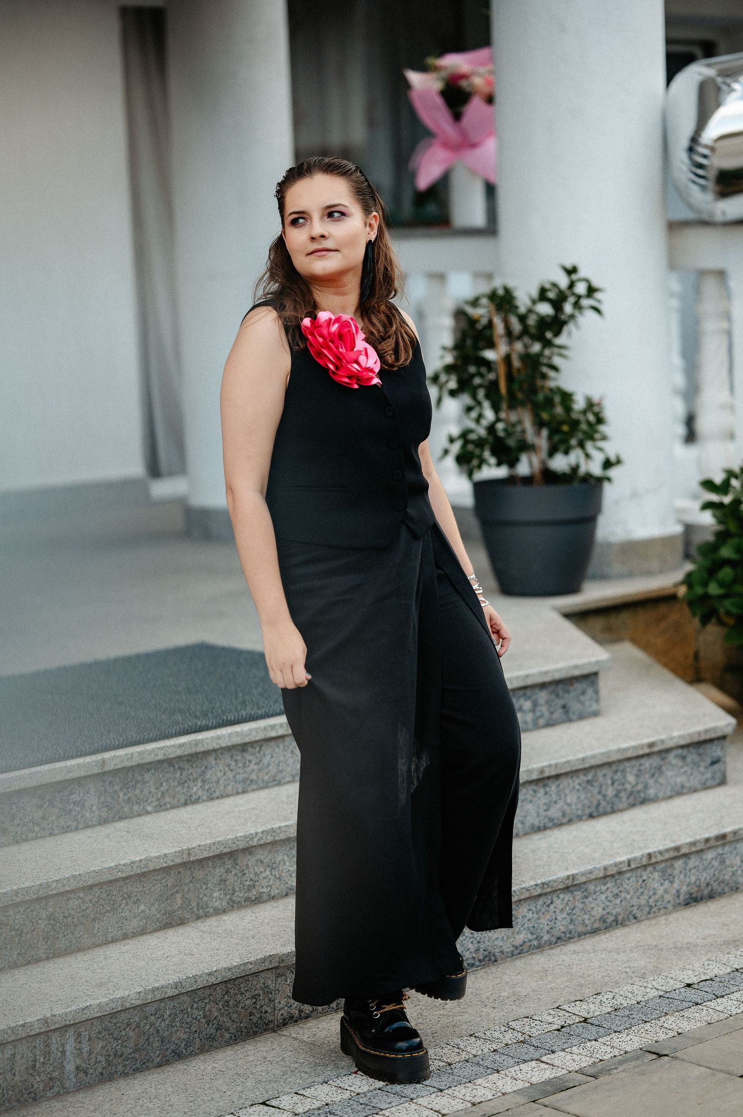 Young woman in black sleeveless maxi dress with red flower corsage posing on outdoor steps.
