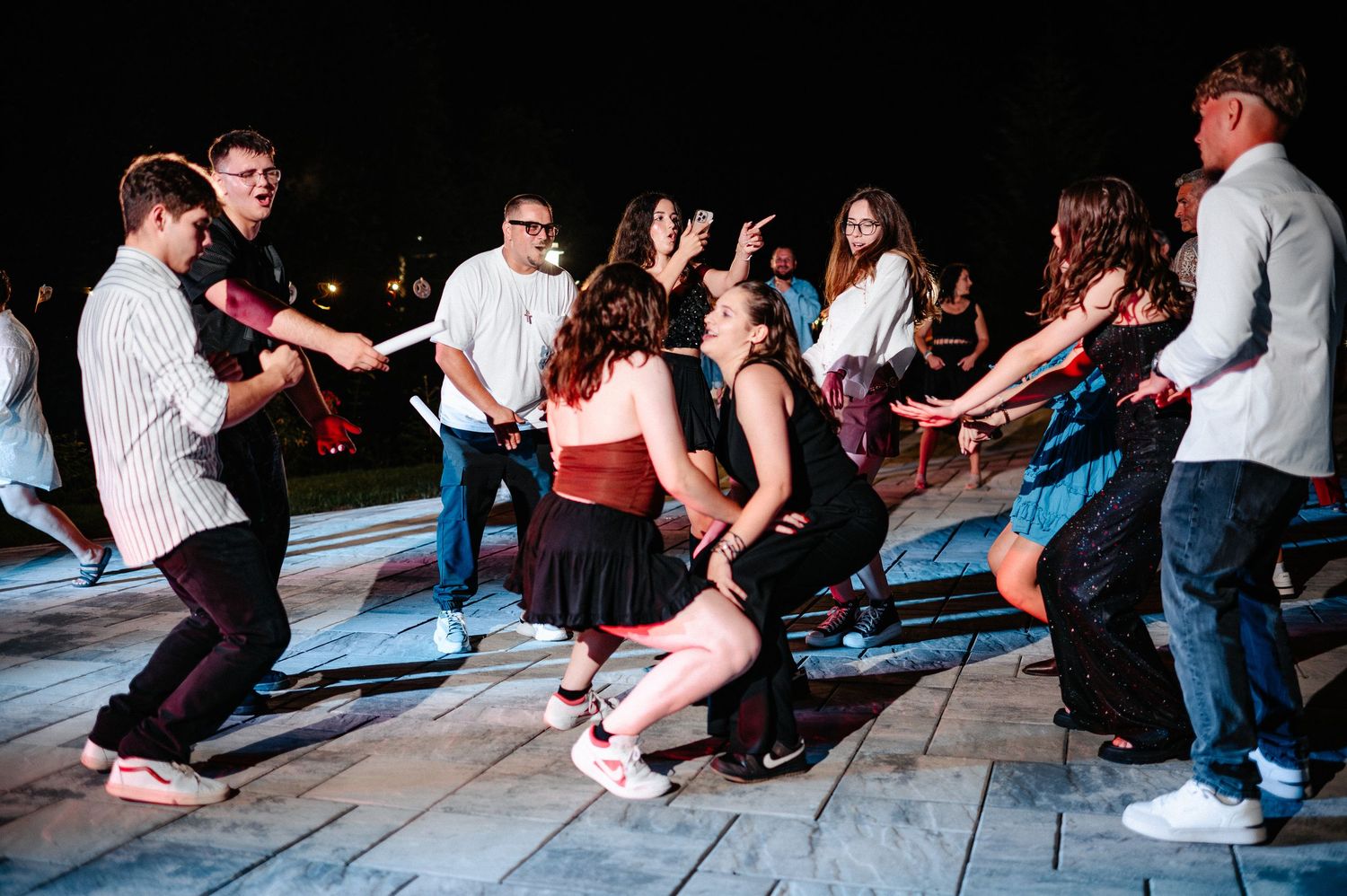 Group of young people dancing energetically at outdoor night party on illuminated dance floor.