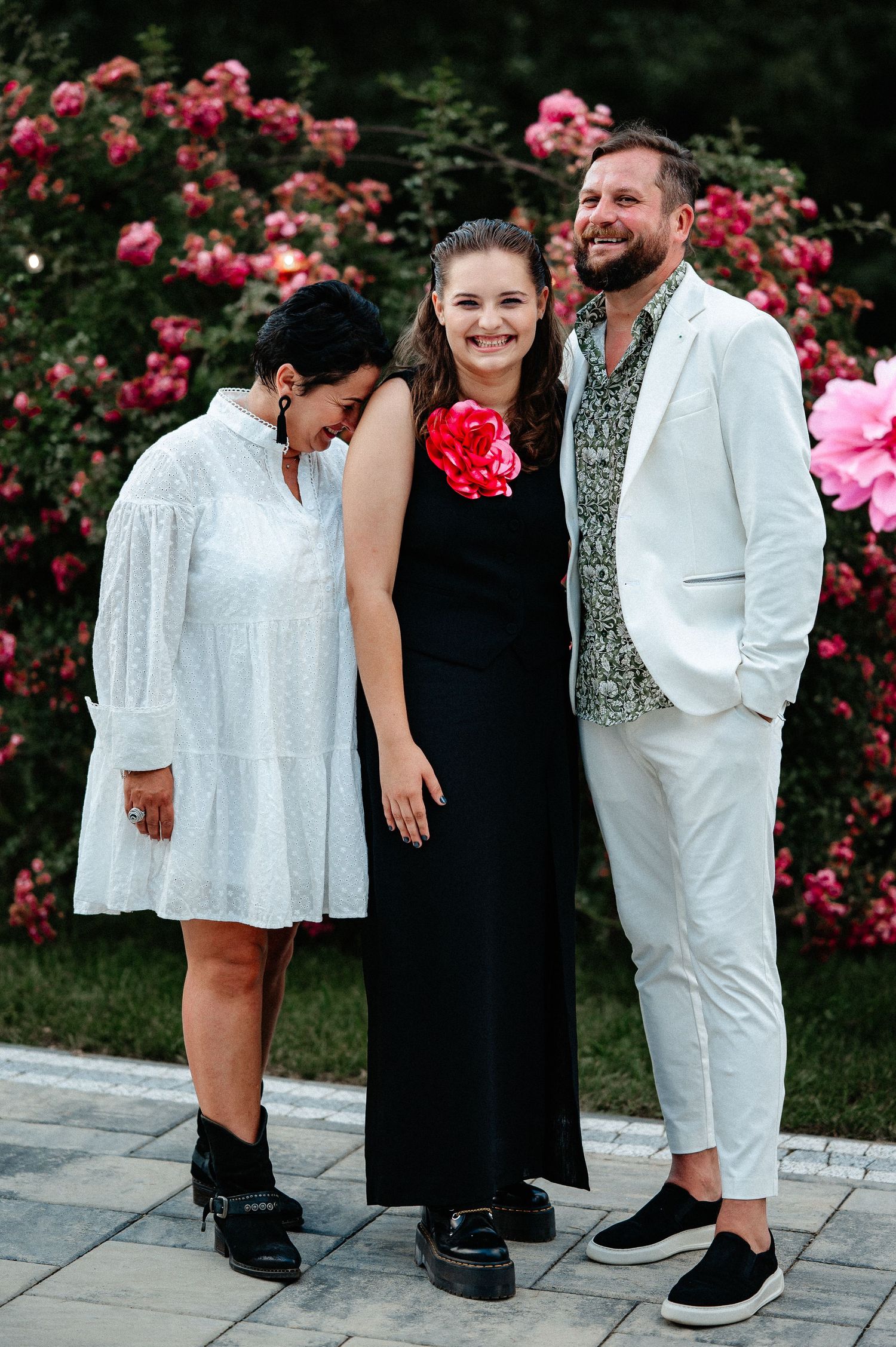 Three people smiling together in front of vibrant pink rose bushes at an outdoor celebration.