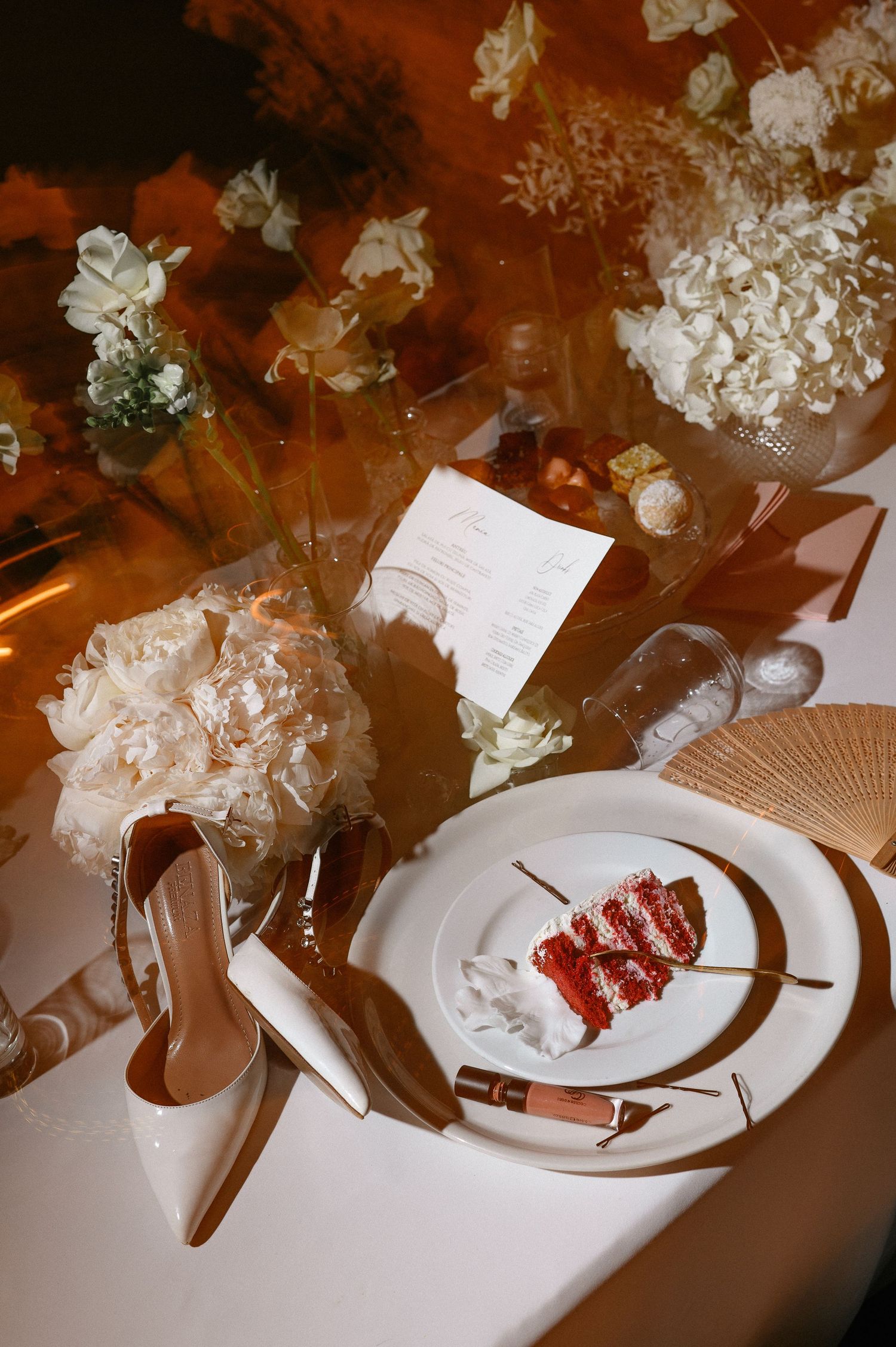 Elegant wedding table setting with red velvet cake slice, white peonies, bridal heels, and gold accents on white linen.