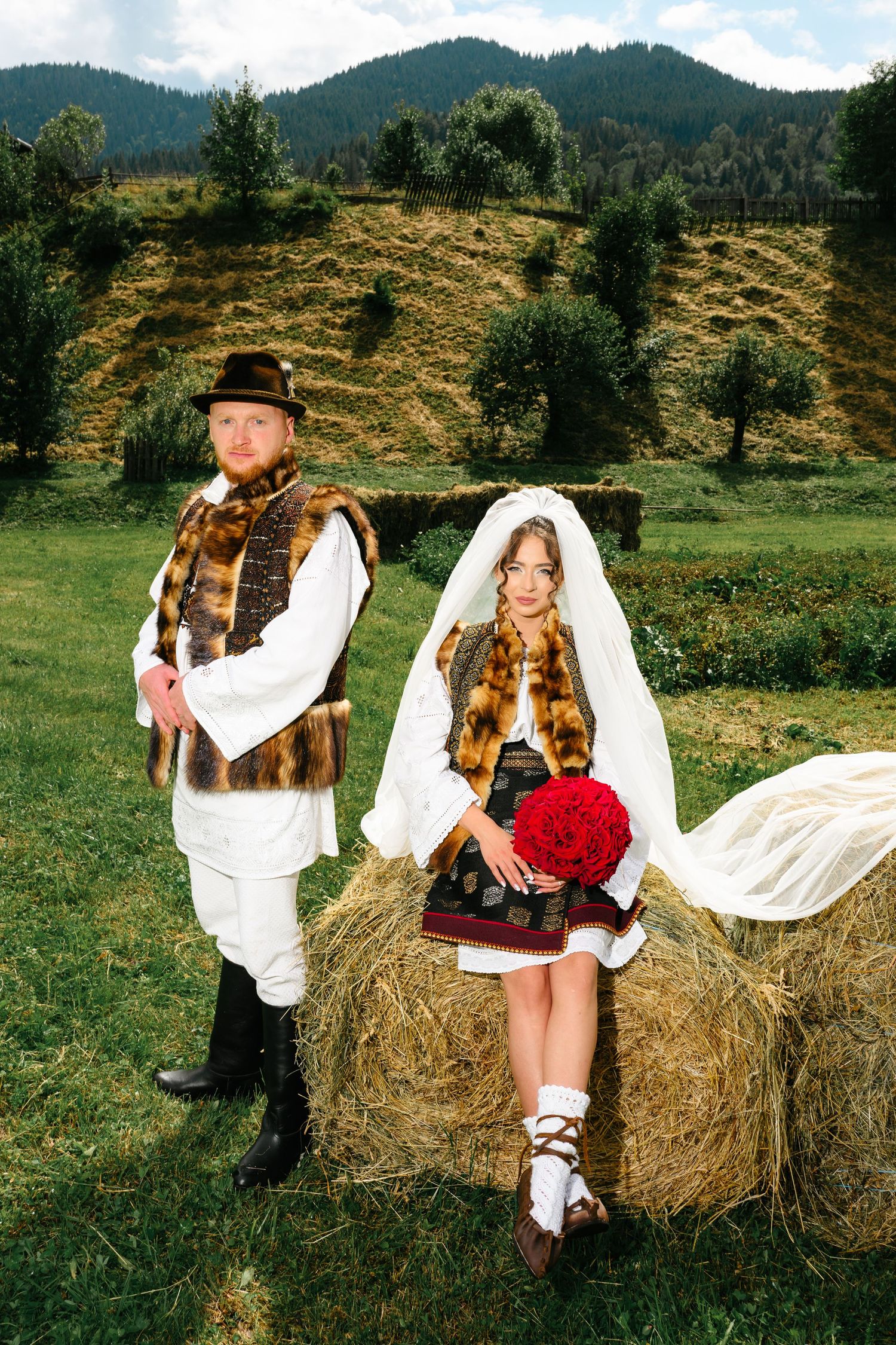 Couple in traditional Romanian folk costumes posing by a hay bale with mountain scenery backdrop.