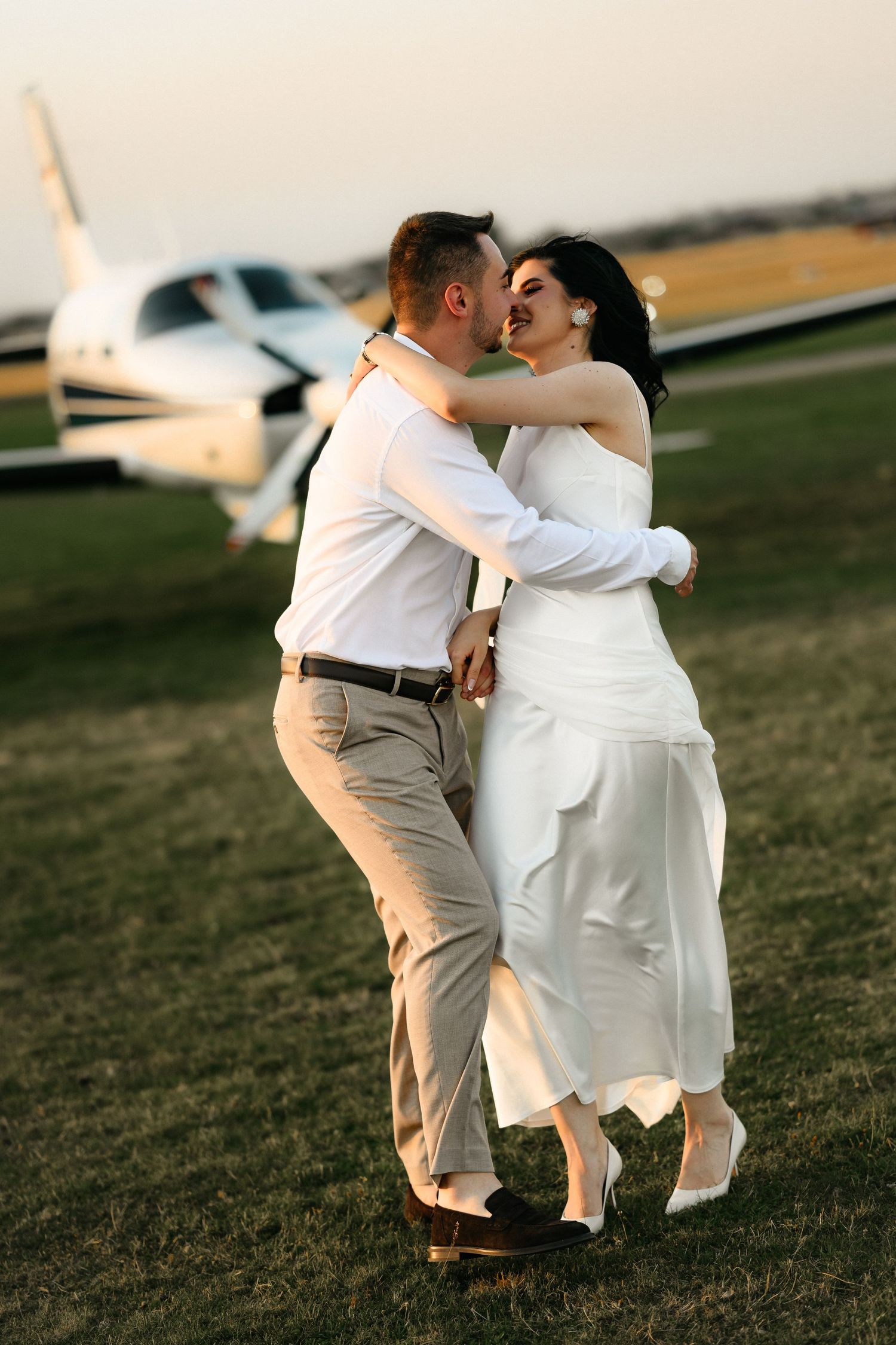 A couple in wedding attire embracing and laughing near a small private plane on a grassy airfield at sunset.