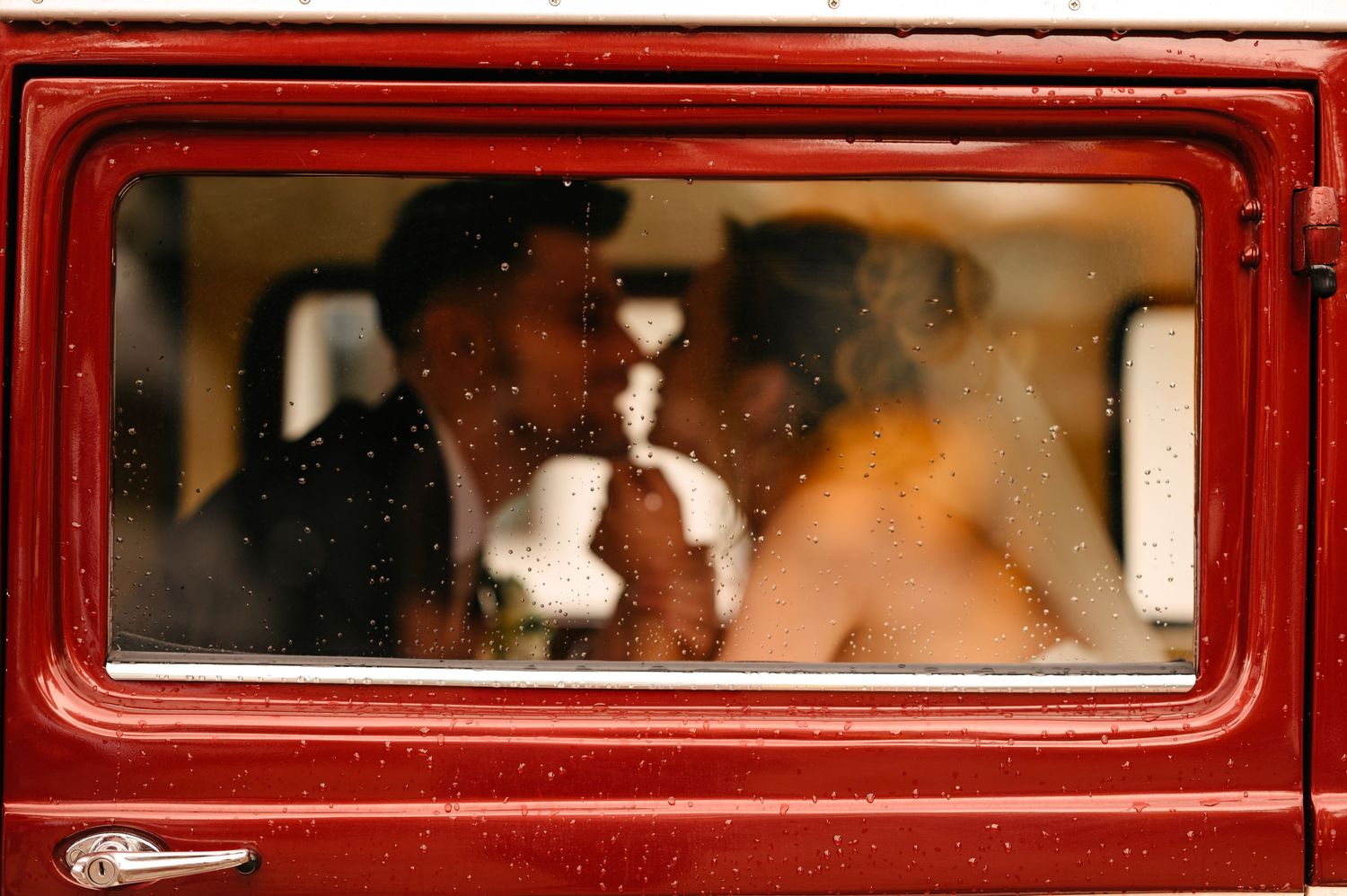 Couple sharing a kiss through a rain-speckled red vintage car window on their wedding day.