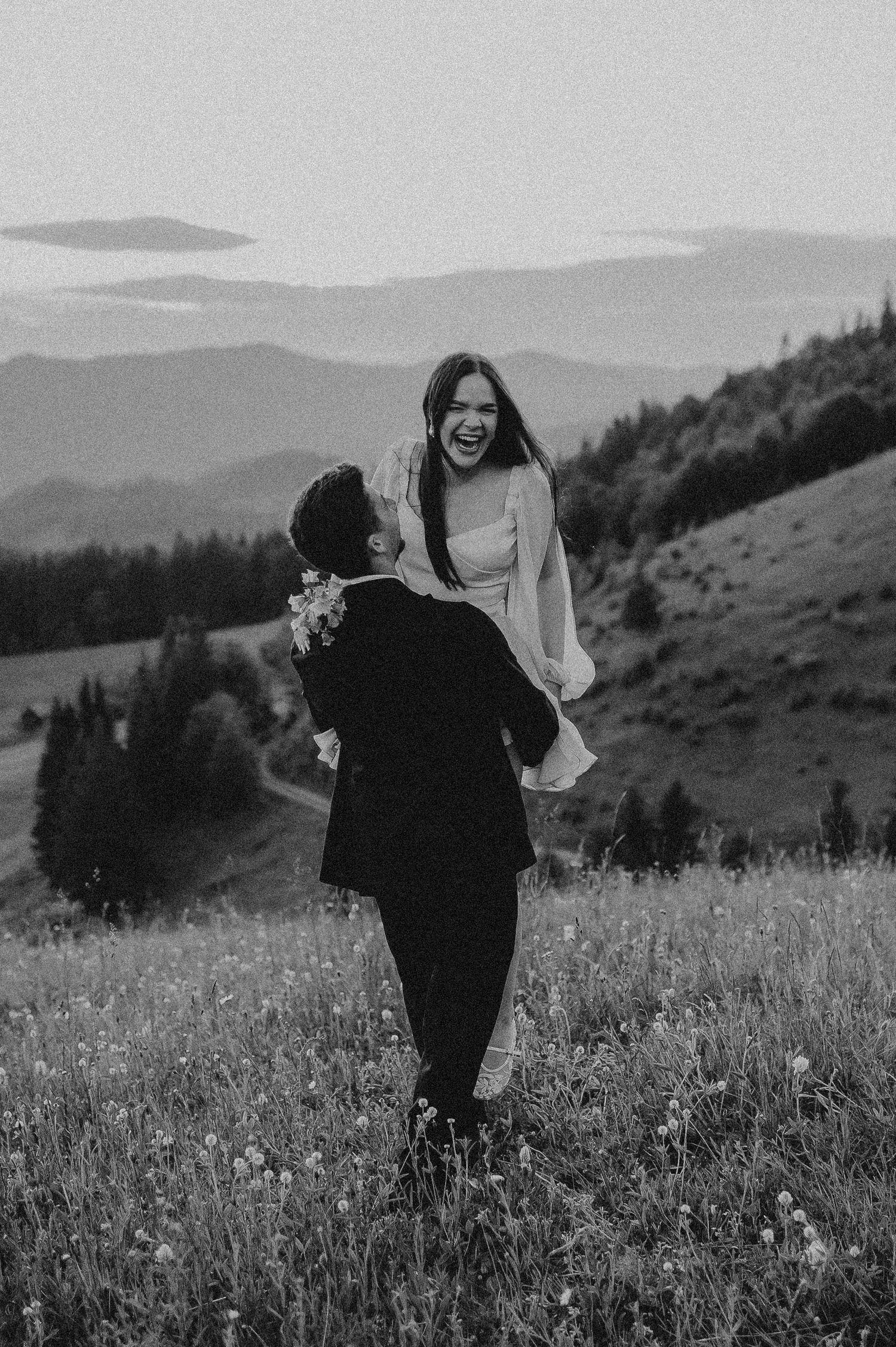 Black and white photo of a groom lifting his laughing bride in a mountain meadow at sunset.