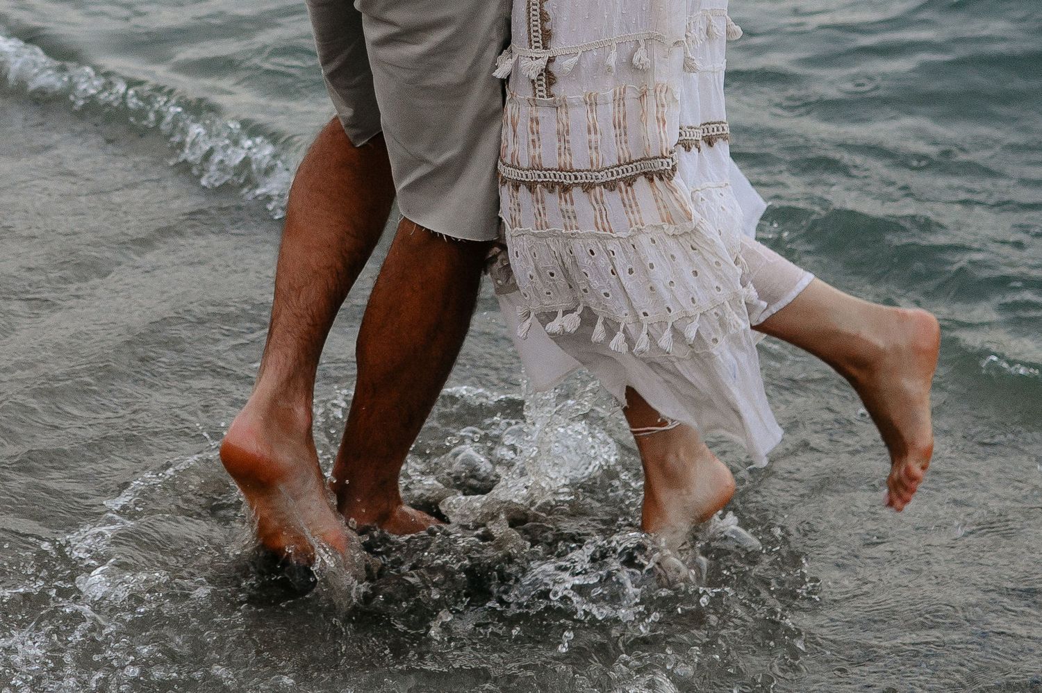 Close-up of a barefoot couple splashing through shallow ocean waves on a sandy beach.
