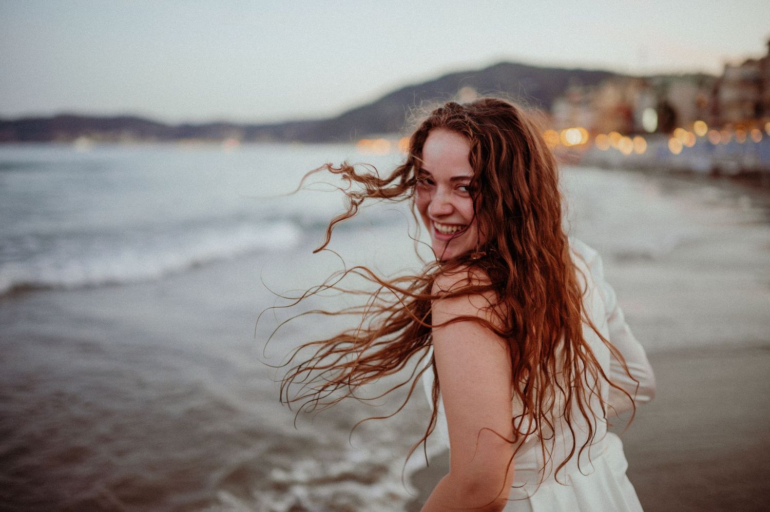 Joyful young woman in white dress smiling on a beach at dusk with wind blowing her long hair.