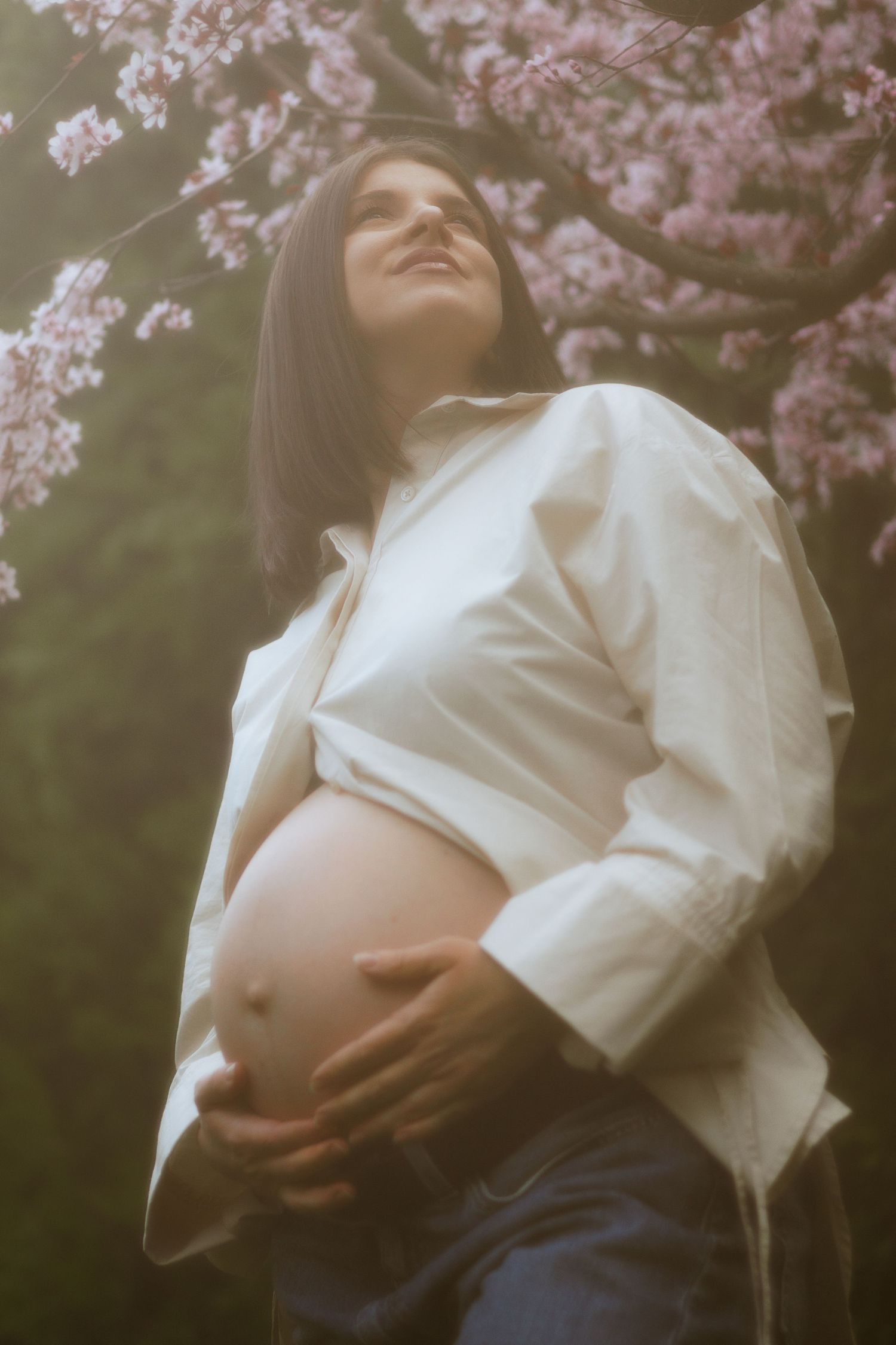 Pregnant woman in white shirt cradling baby bump under pink cherry blossom tree in soft light.