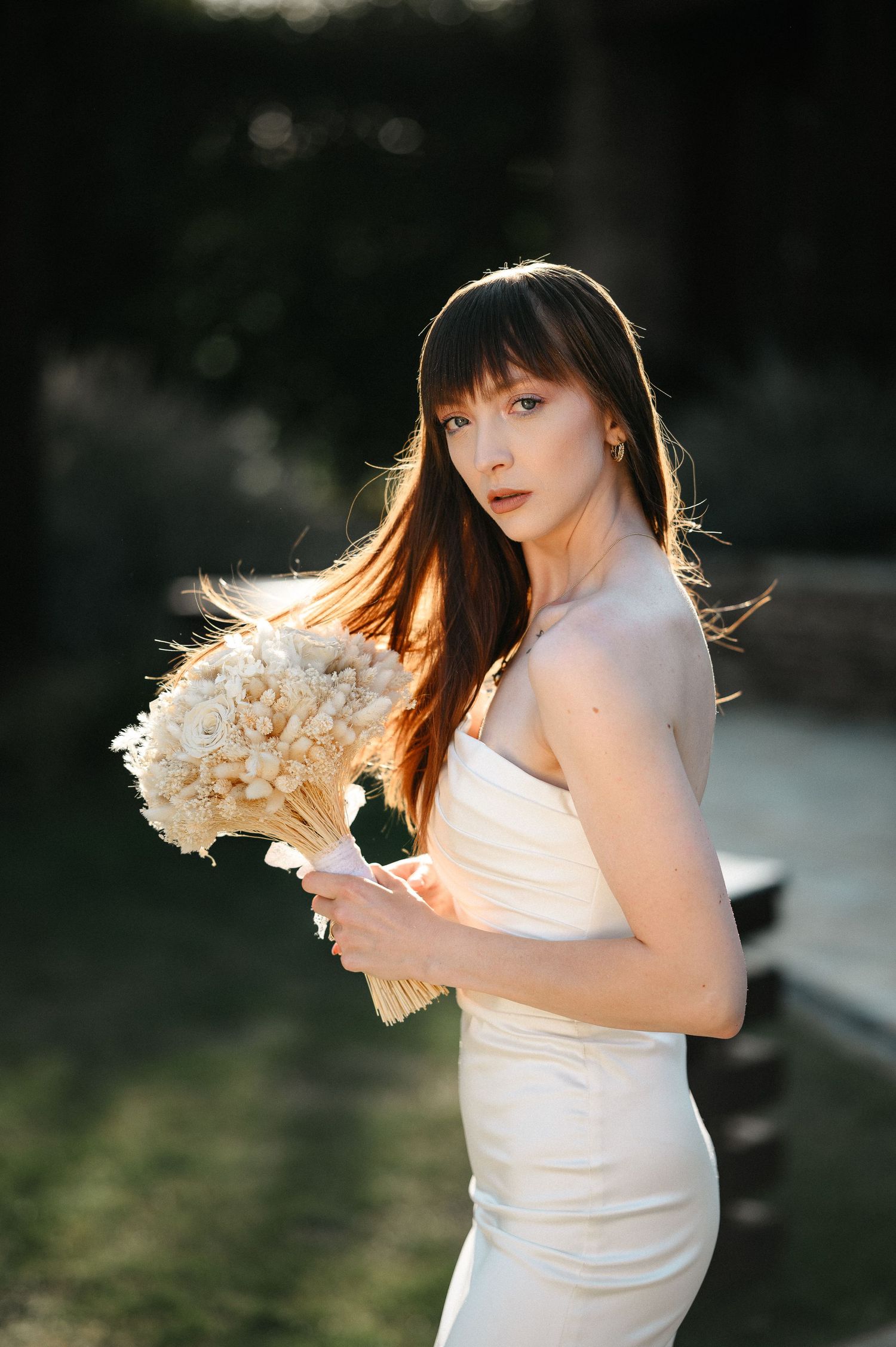 Elegant bride with bangs in white strapless gown holds dried flower bouquet in golden garden light.